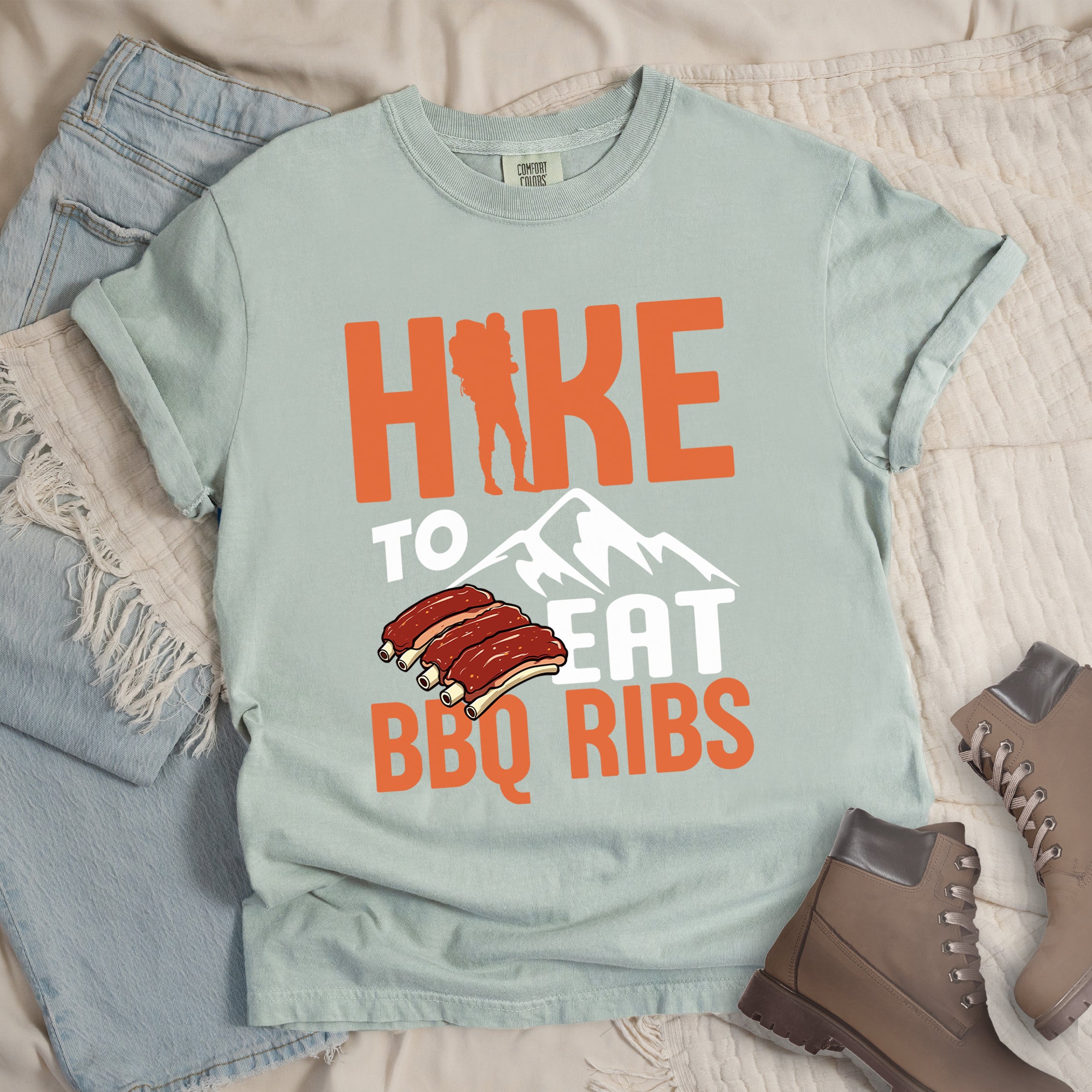 Bay (Light Gray) t-shirt with text "HIKE TO EAT BBQ RIBS". The word "HIKE" appears in orange with a hiker silhouette replacing the "I". Below that is "TO" in white text above a white mountain graphic. "EAT" appears in white, and "BBQ RIBS" in orange at the bottom. Between "EAT" and "BBQ RIBS" is an illustration of a rack of barbecue ribs with exposed bones.