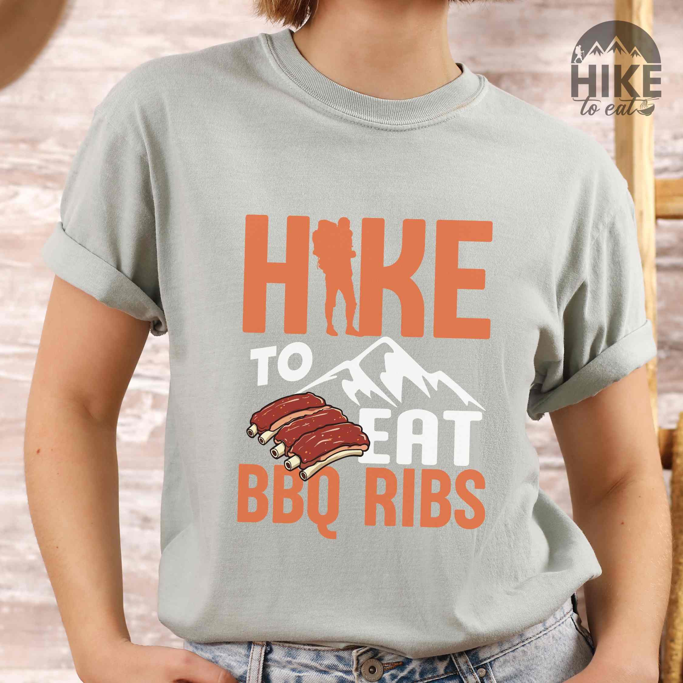 Woman wearing bay "Hike to eat BBQ ribs" colored vintage tee with bold typography, mountains, juicy grilled bbq ribs illustration, and backpacker for bbq enthusiasts