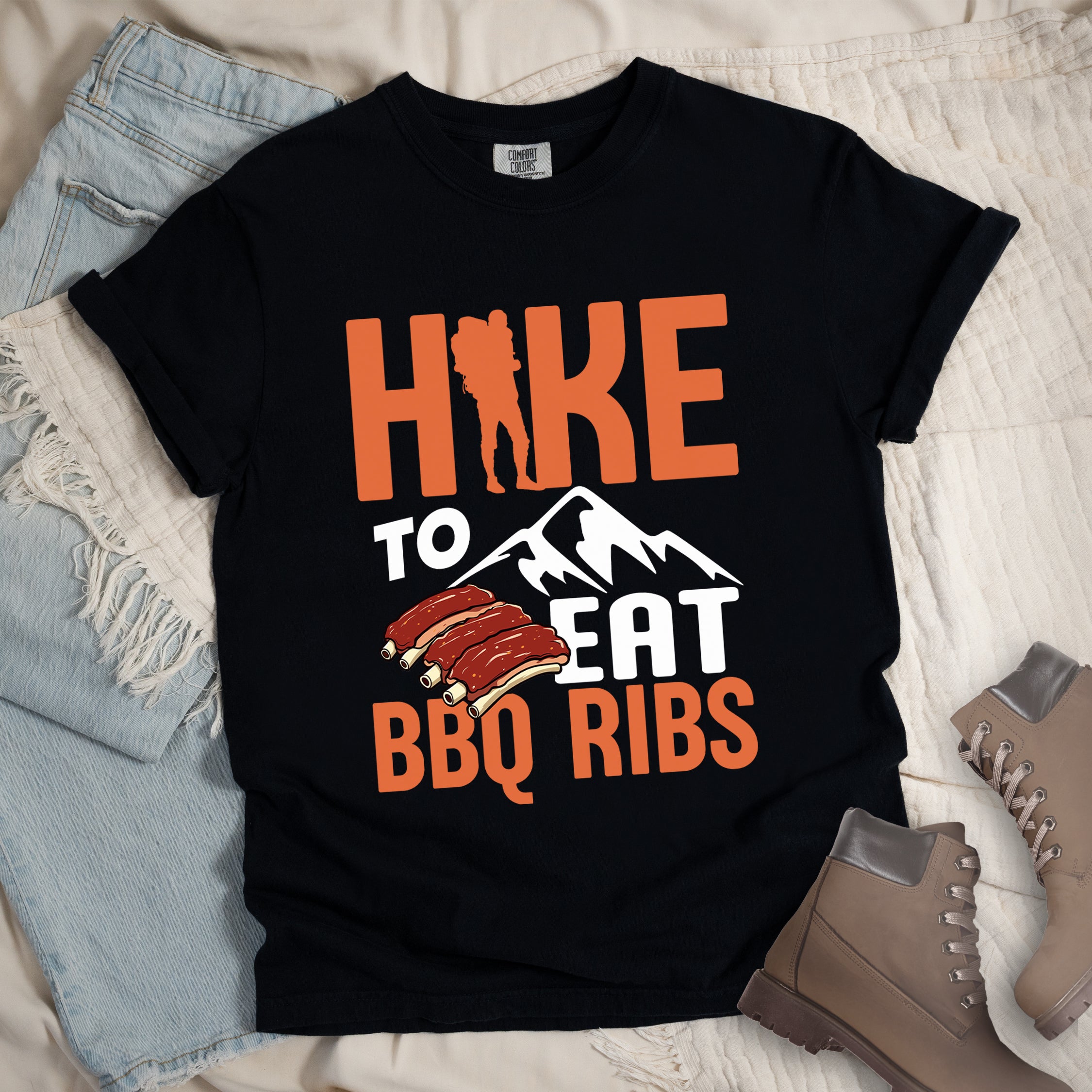 Black t-shirt with text "HIKE TO EAT BBQ RIBS". The word "HIKE" appears in orange with a hiker silhouette replacing the "I". Below that is "TO" in white text above a white mountain graphic. "EAT" appears in white, and "BBQ RIBS" in orange at the bottom. Between "EAT" and "BBQ RIBS" is an illustration of a rack of barbecue ribs with exposed bones.