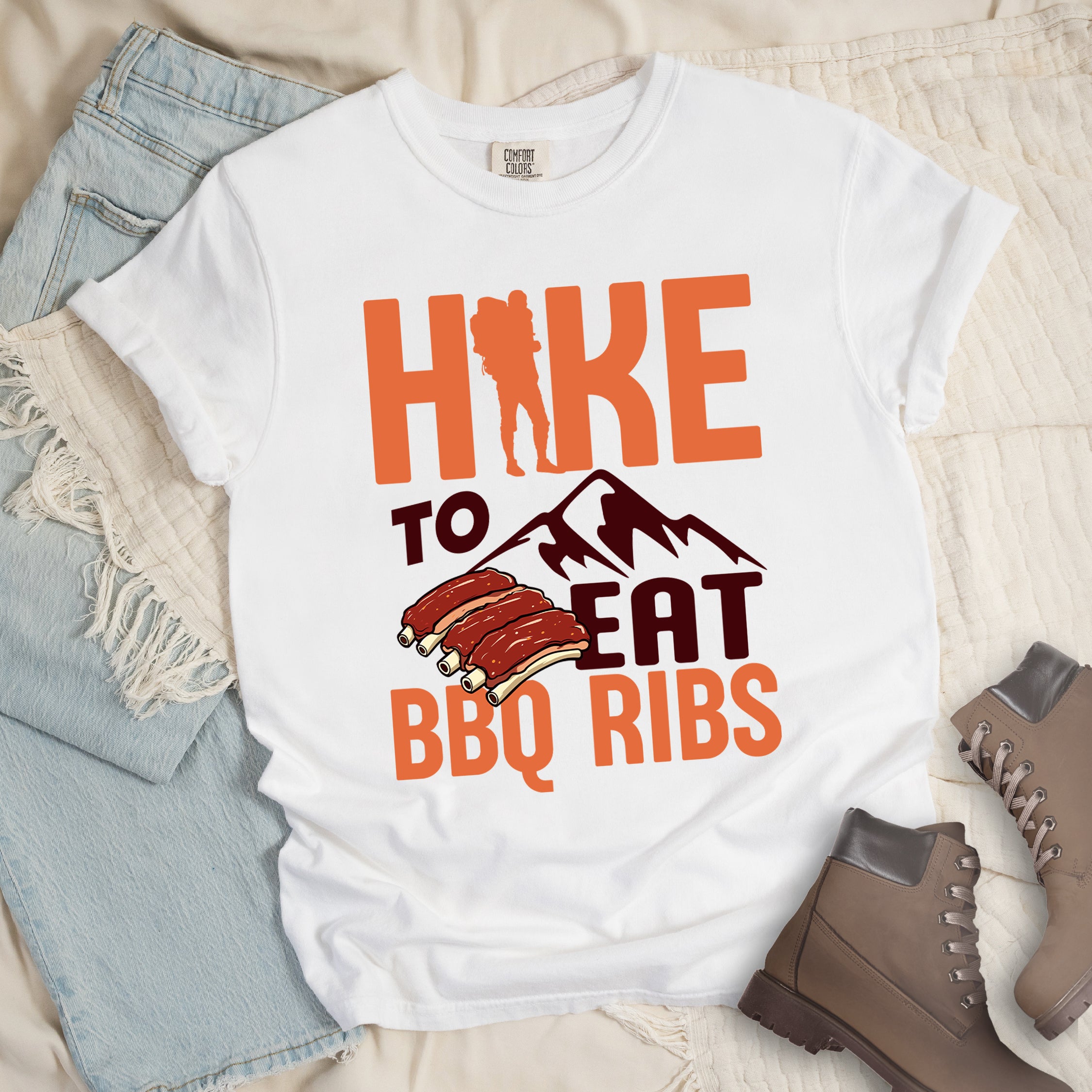 White t-shirt with text "HIKE TO EAT BBQ RIBS". The word "HIKE" appears in orange with a hiker silhouette replacing the "I". Below that is "TO" in deep brown-red text above a deep brown-red mountain graphic. "EAT" appears in deep brown-red, and "BBQ RIBS" in orange at the bottom. Between "EAT" and "BBQ RIBS" is an illustration of a rack of barbecue ribs with exposed bones.