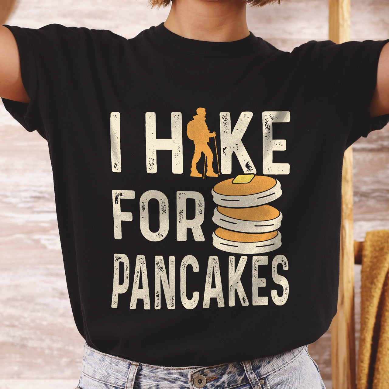 Woman wearing black colored Pancake breakfast lover vintage tee I Hike for Pancakes with distressed cream typography character silhouette with trekking pole and illustrated stacked pancakes with butter on Comfort Colors shirt for brunch and food enthusiasts, hikers and foodies.