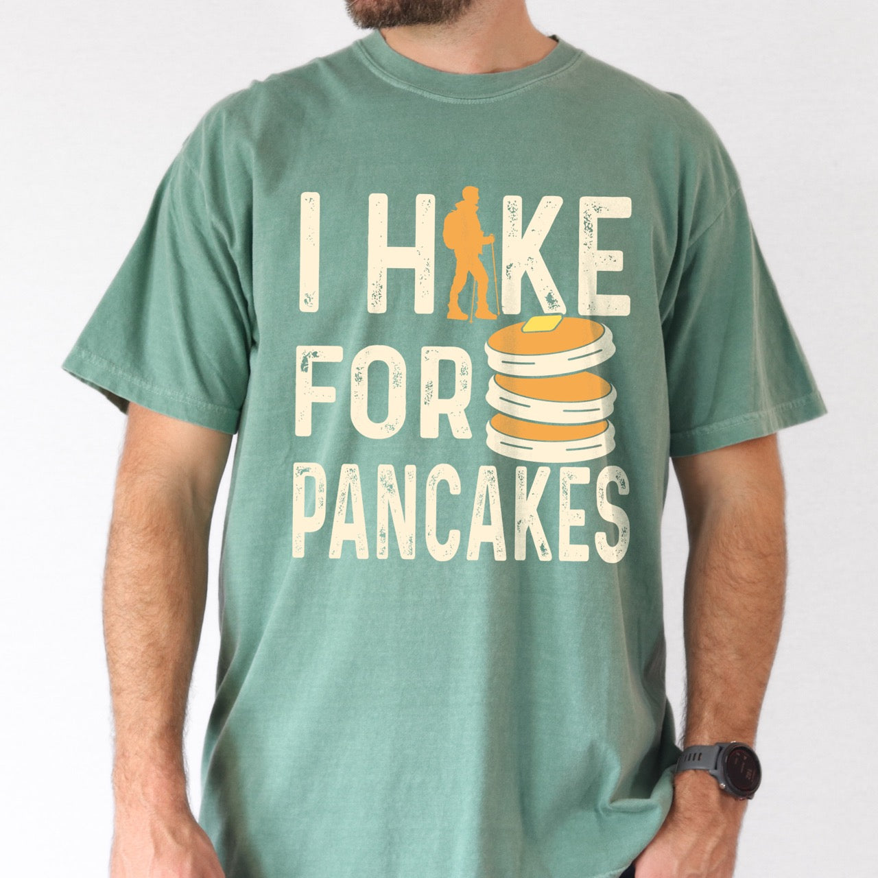 Man wearing light green colored Pancake breakfast lover vintage tee I Hike for Pancakes with distressed cream typography character silhouette with trekking pole and illustrated stacked pancakes with butter on Comfort Colors shirt for brunch and food enthusiasts, hikers and foodies.