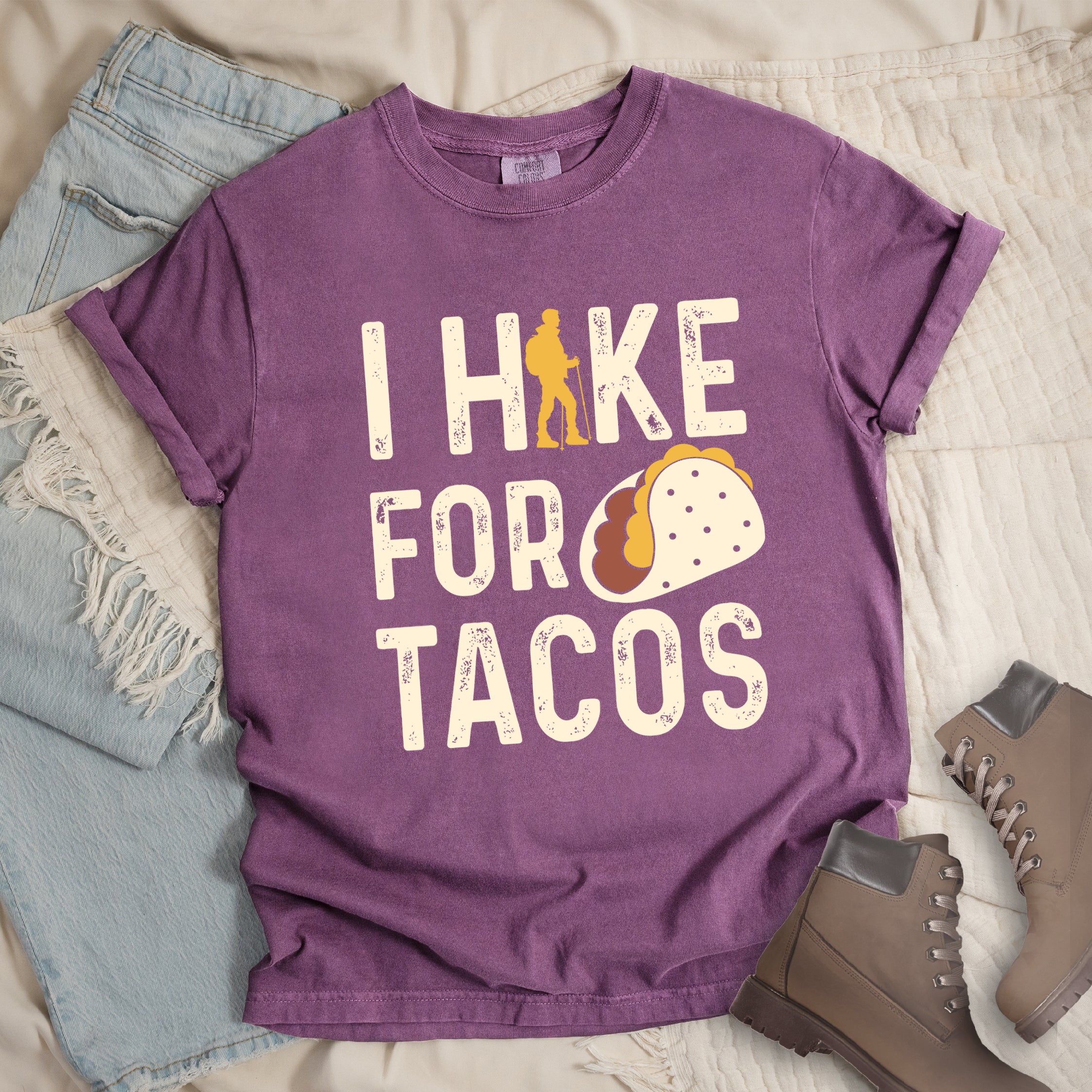 Berry (Dark Violet) graphic t-shirt with 'I Hike for Tacos' text in cream lettering featuring orange hiker silhouette and colorful taco illustration, styled with light wash jeans and brown hiking boots.