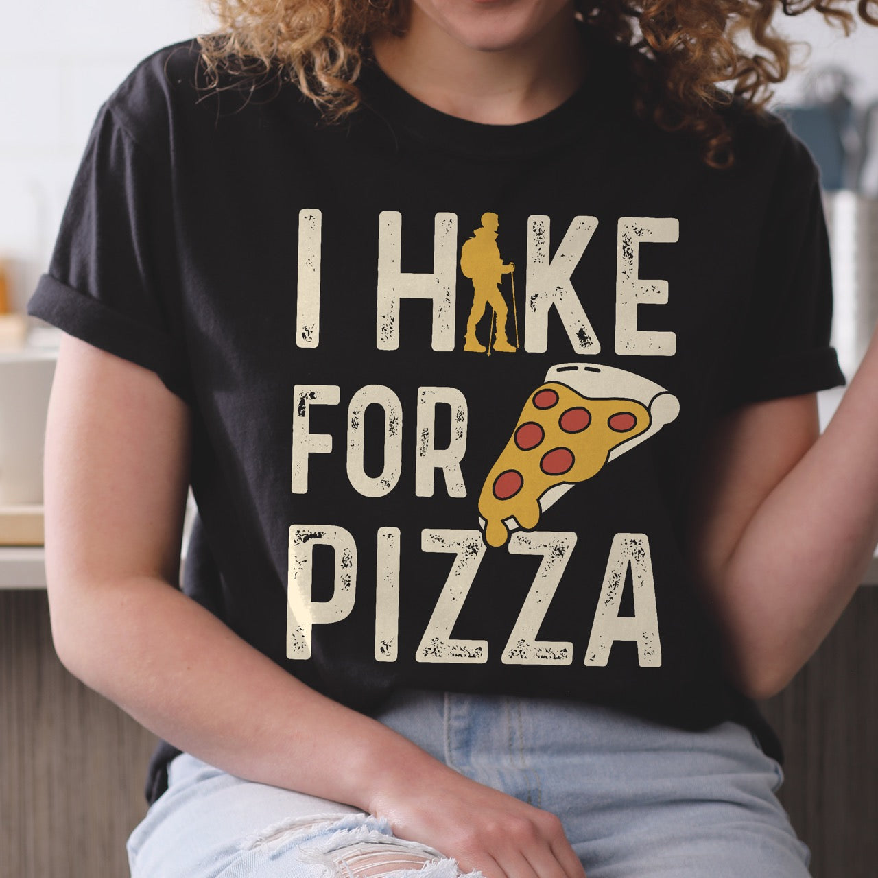 Woman wearing I Hike for Pizza graphic tee with distressed cream typography, pepperoni pizza slice illustration, and gold hiker silhouette on Comfort Colors shirt for Italian food enthusiasts and mountain trail lovers in black