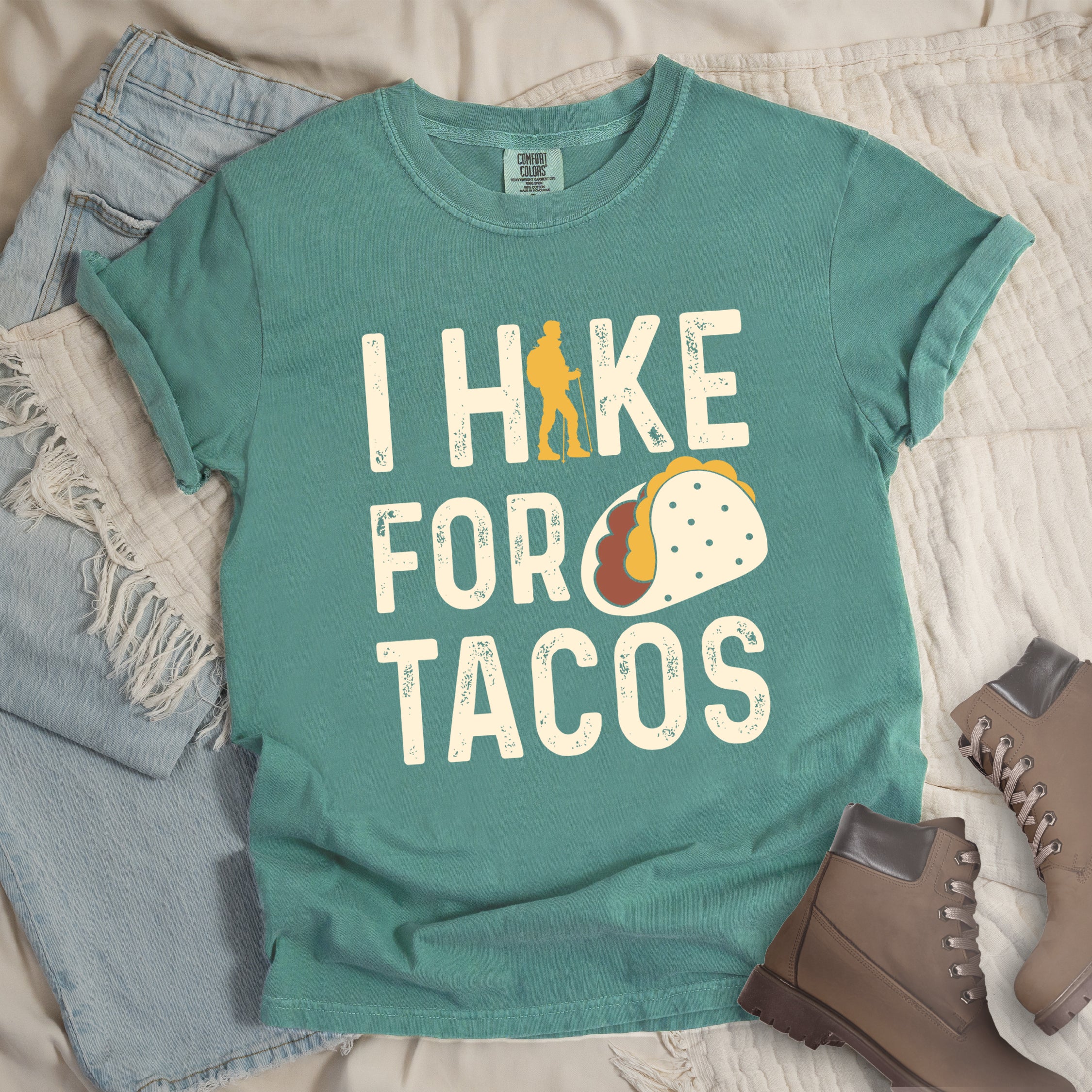 Light Green graphic t-shirt with 'I Hike for Tacos' text in cream lettering featuring orange hiker silhouette and colorful taco illustration, styled with light wash jeans and brown hiking boots
