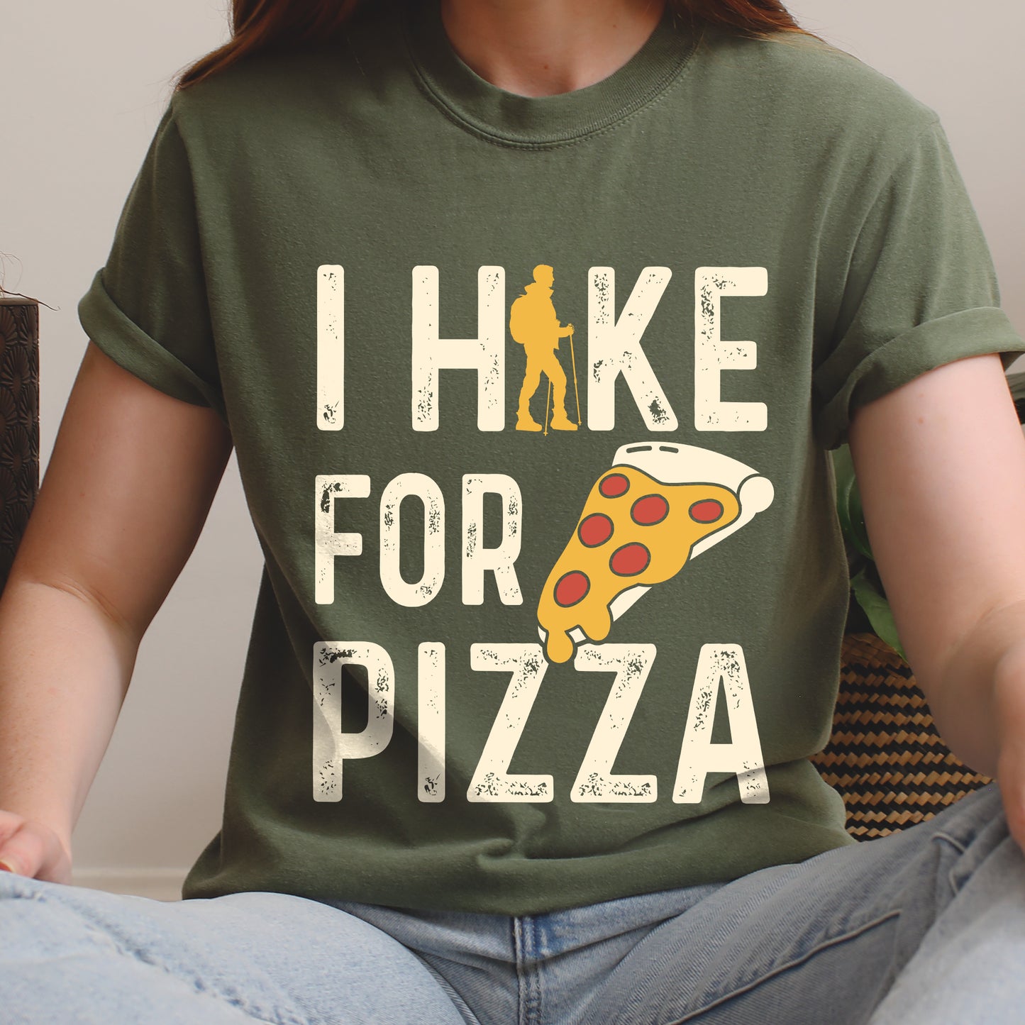 Moss (Dark Green) graphic t-shirt with 'I Hike for Pizza' text in cream lettering featuring orange hiker silhouette and pizza slice illustration with pepperoni toppings.
