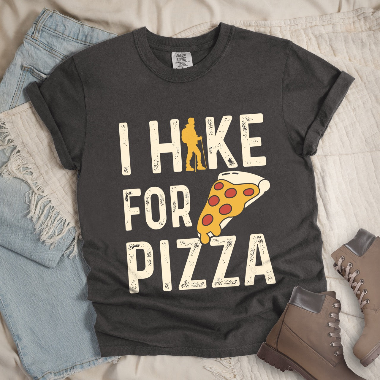 pepper  I Hike for Pizza graphic tee with distressed cream typography, pepperoni pizza slice illustration, and gold hiker silhouette on Comfort Colors shirt for Italian food enthusiasts and mountain trail lovers layed flat with boots and jeans