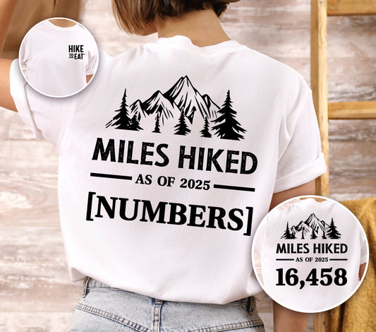 White Hike to Eat t-shirt front and back view showing small black chest detail and large back design featuring 'Milestone Tracker Personalized' with mountain and pine tree silhouettes, 'Miles Hiked as of 2025' text and customizable numbers field, shown with close-up detail displaying example number 16,458.