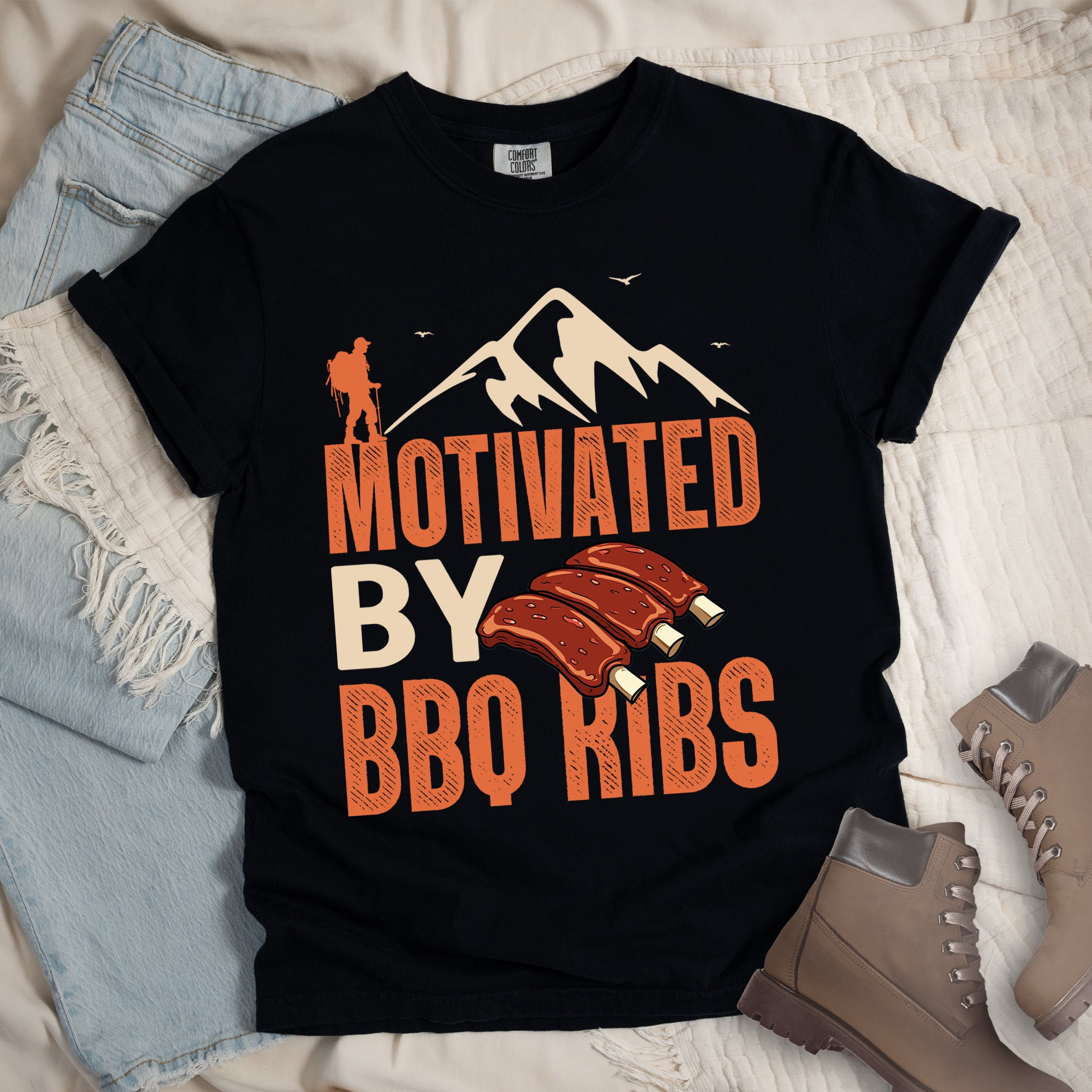 Black t-shirt with text "MOTIVATED BY BBQ RIBS". At the top is a cream-colored mountain silhouette with flying birds and an orange hiker figure. "MOTIVATED" appears in large orange textured letters, "BY" in cream/tan, and "BBQ RIBS" in matching orange. Next to "BY" and above "BBQ RIBS" is an illustration of three barbecue ribs with exposed bones. The design combines hiking and food themes.