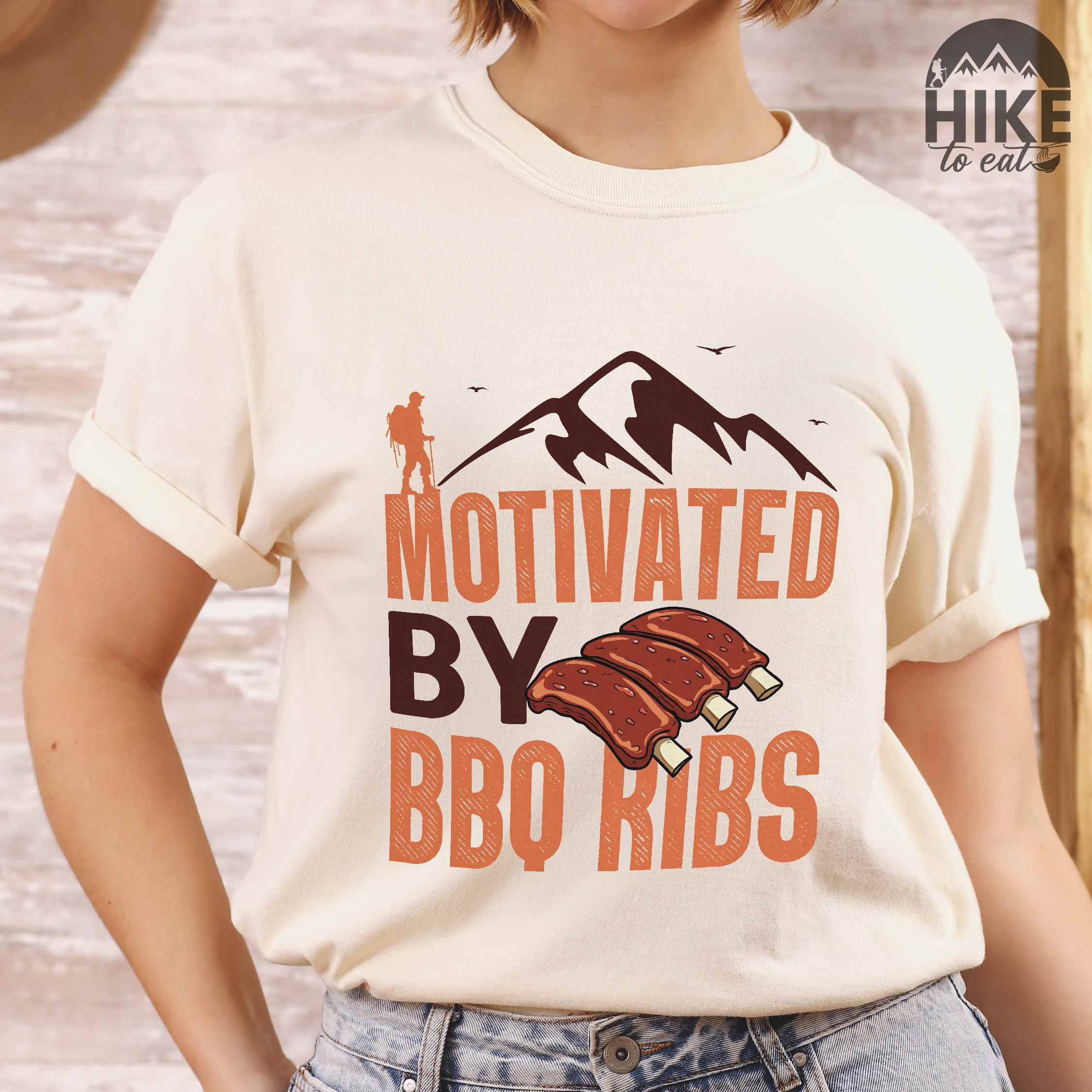 Woman wearing ivory "Motivated by BBQ ribs" colored vintage tee with bold typography, mountains, juicy grilled bbq ribs illustration, and backpacker for bbq enthusiasts