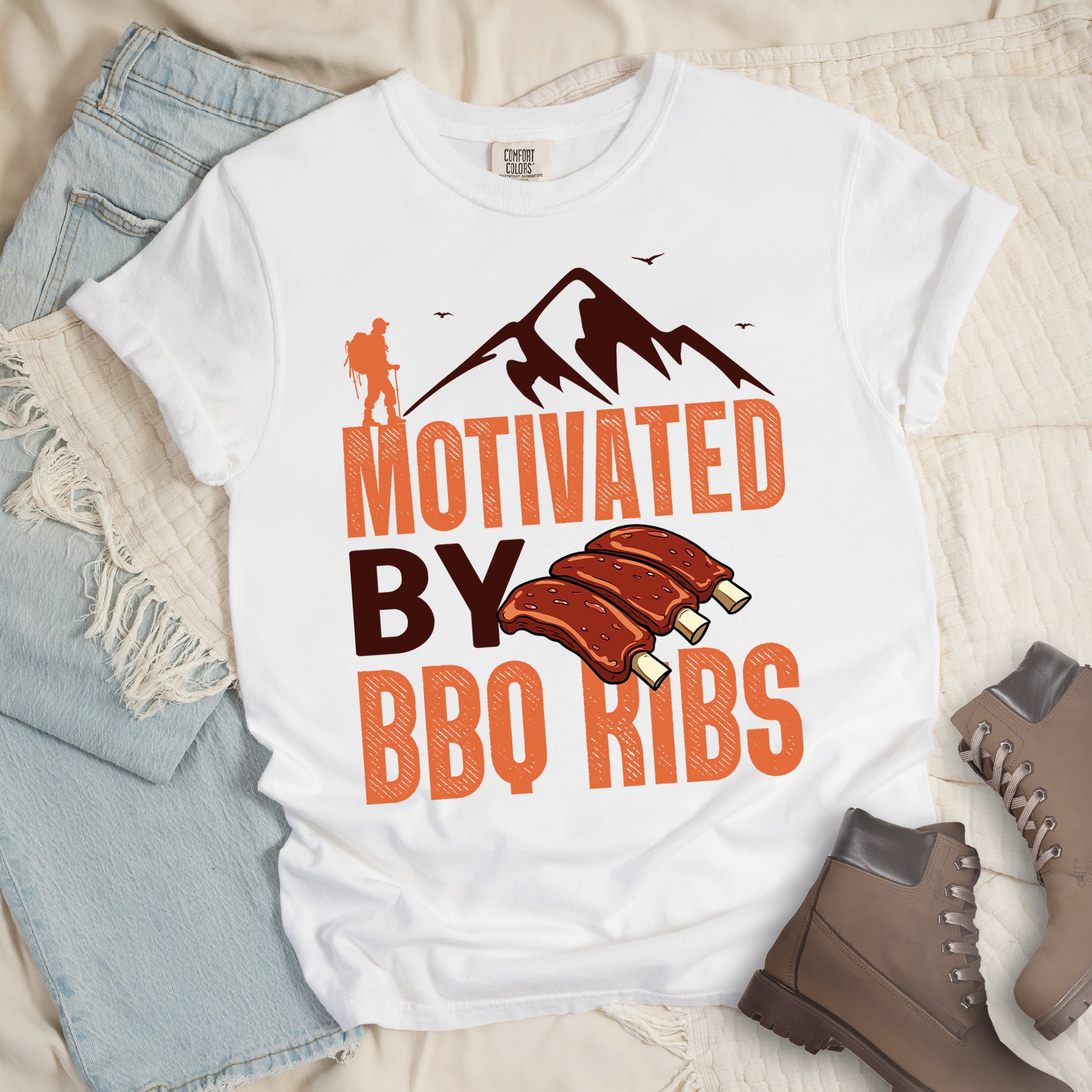 White t-shirt with text "MOTIVATED BY BBQ RIBS". At the top is a deep brown-colored mountain silhouette with flying birds and an orange hiker figure. "MOTIVATED" appears in large orange textured letters, "BY" in deep brown, and "BBQ RIBS" in matching orange. Next to "BY" and above "BBQ RIBS" is an illustration of three barbecue ribs with exposed bones. The design combines hiking and food themes.