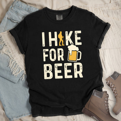 Black graphic t-shirt with 'I Hike for Beer' text in cream lettering featuring orange hiker silhouette and beer mug illustration with foam, styled with light wash jeans and brown hiking boots.
