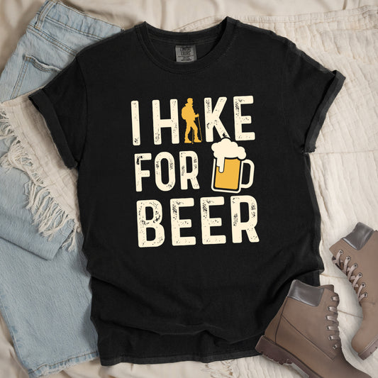 Black graphic t-shirt with 'I Hike for Beer' text in cream lettering featuring orange hiker silhouette and beer mug illustration with foam, styled with light wash jeans and brown hiking boots.