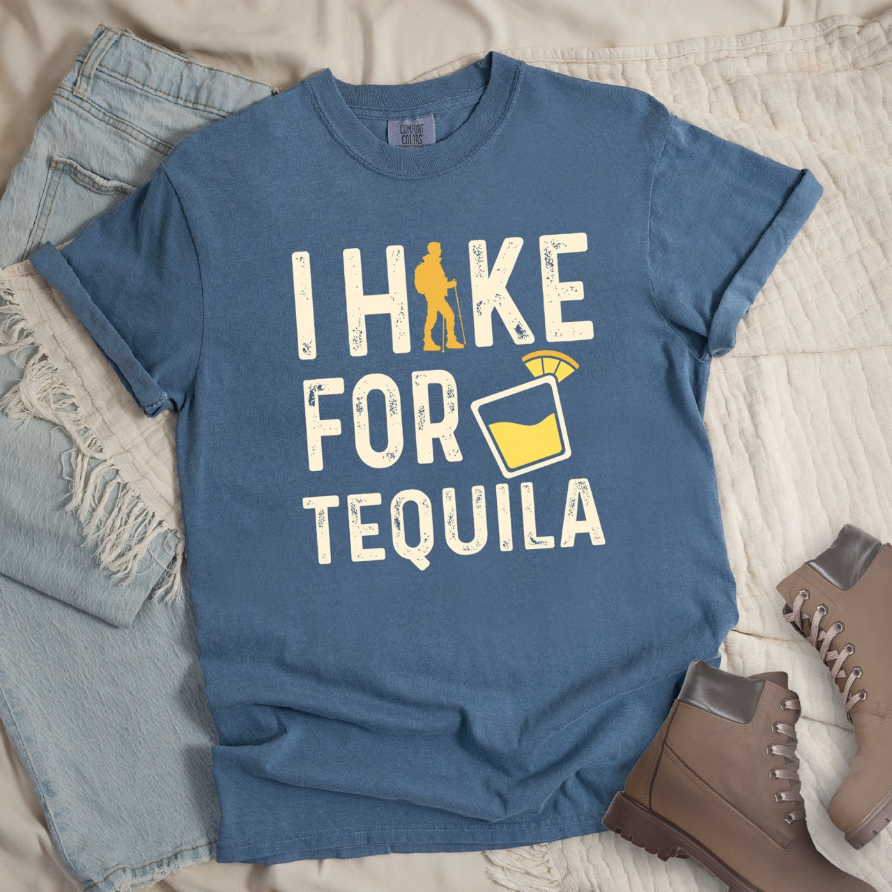 Blue Jean (blue) Tequila margarita lover statement tee I Hike for Tequila with vintage distressed cream typography character silhouette with trekking pole and illustrated tilted margarita glass with lime garnish on Comfort Colors shirt for drink enthusiasts for hikers and foodies - laid flat with boots adn pants 
