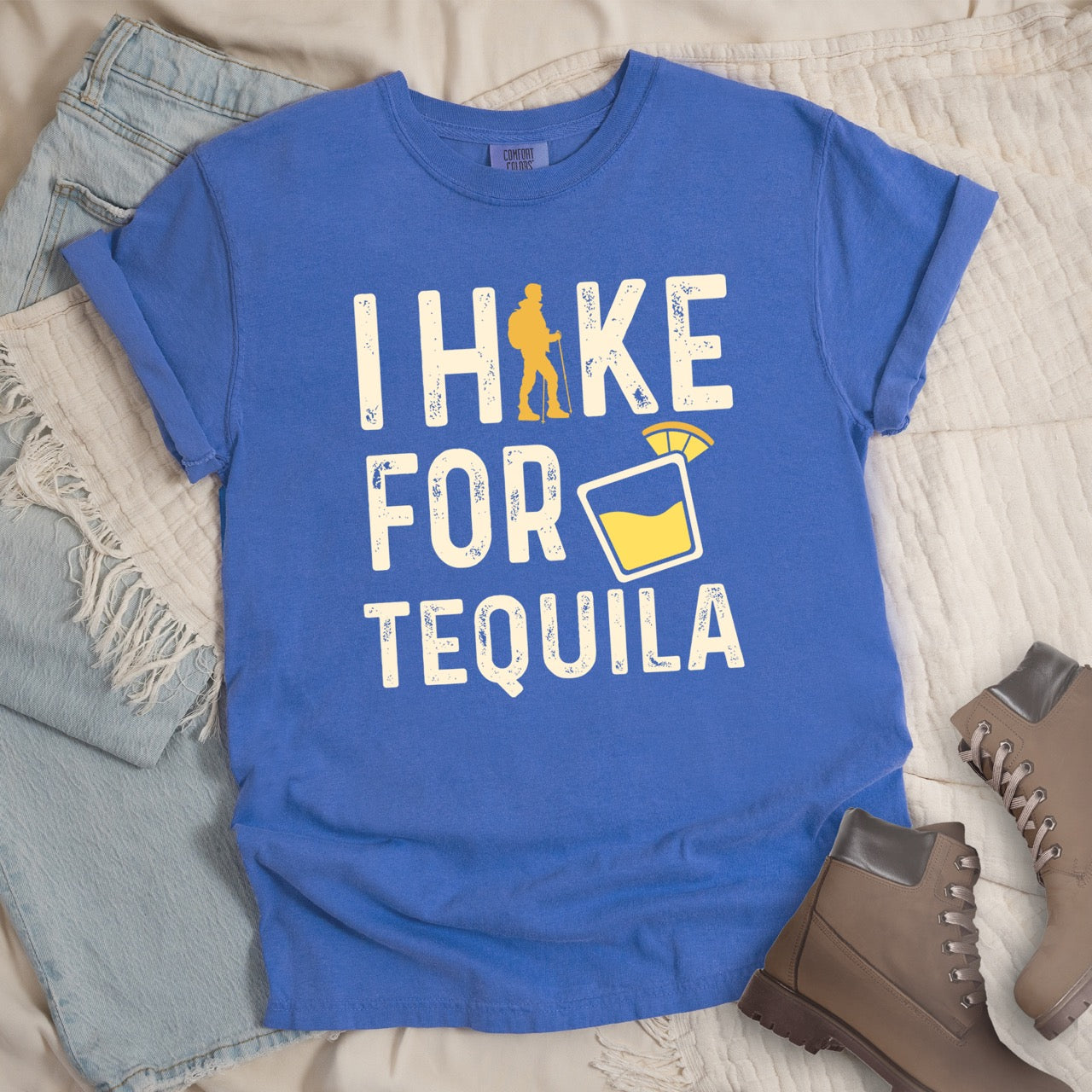 Flo Blue (Bright bluish purple) Tequila margarita lover statement tee I Hike for Tequila with vintage distressed cream typography character silhouette with trekking pole and illustrated tilted margarita glass with lime garnish on Comfort Colors shirt for drink enthusiasts for hikers and foodies - laid flat with pants and boots. 