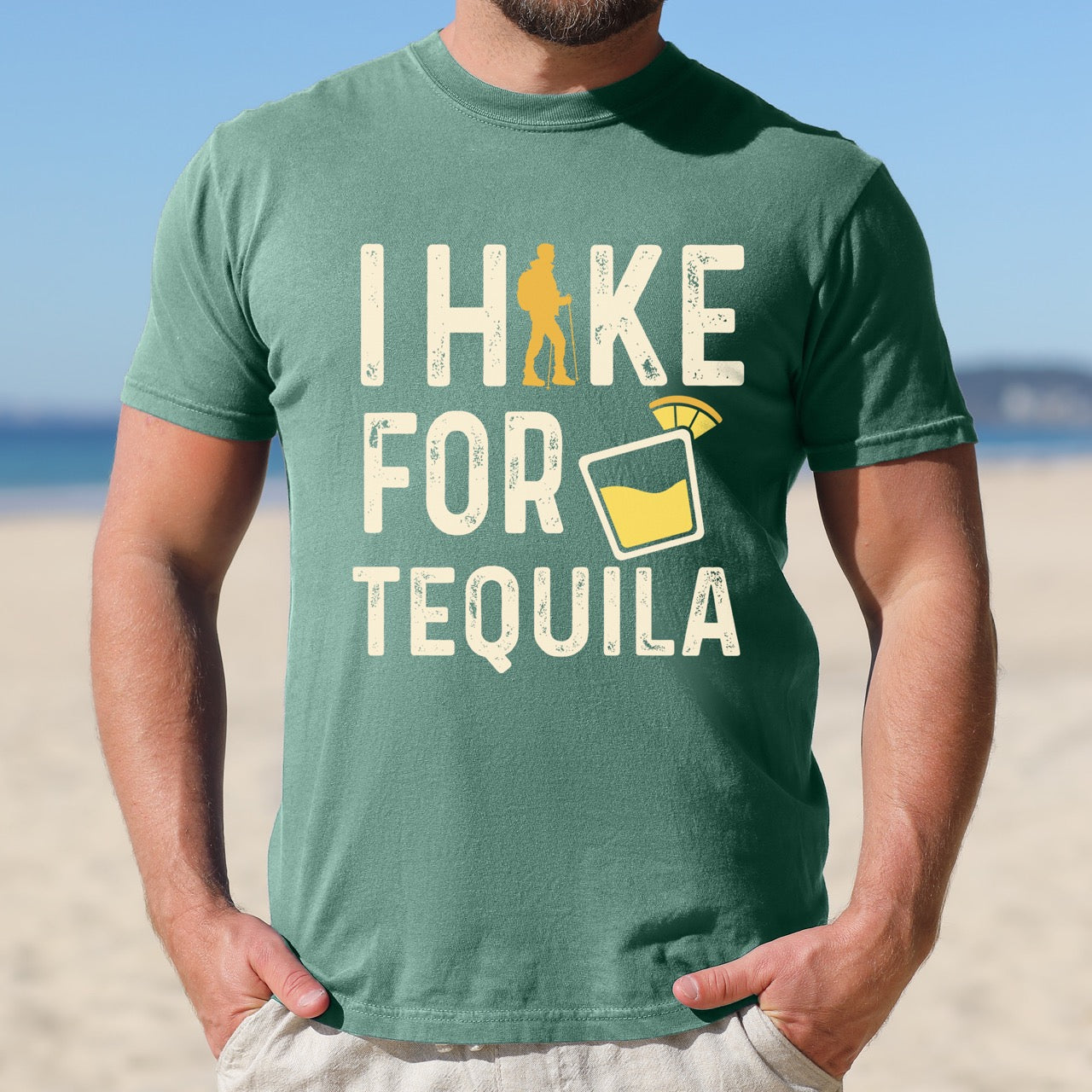 Man wearing light green Tequila margarita lover statement tee I Hike for Tequila with vintage distressed cream typography character silhouette with trekking pole and illustrated tilted margarita glass with lime garnish on Comfort Colors shirt for drink enthusiasts for hikers and foodies. 