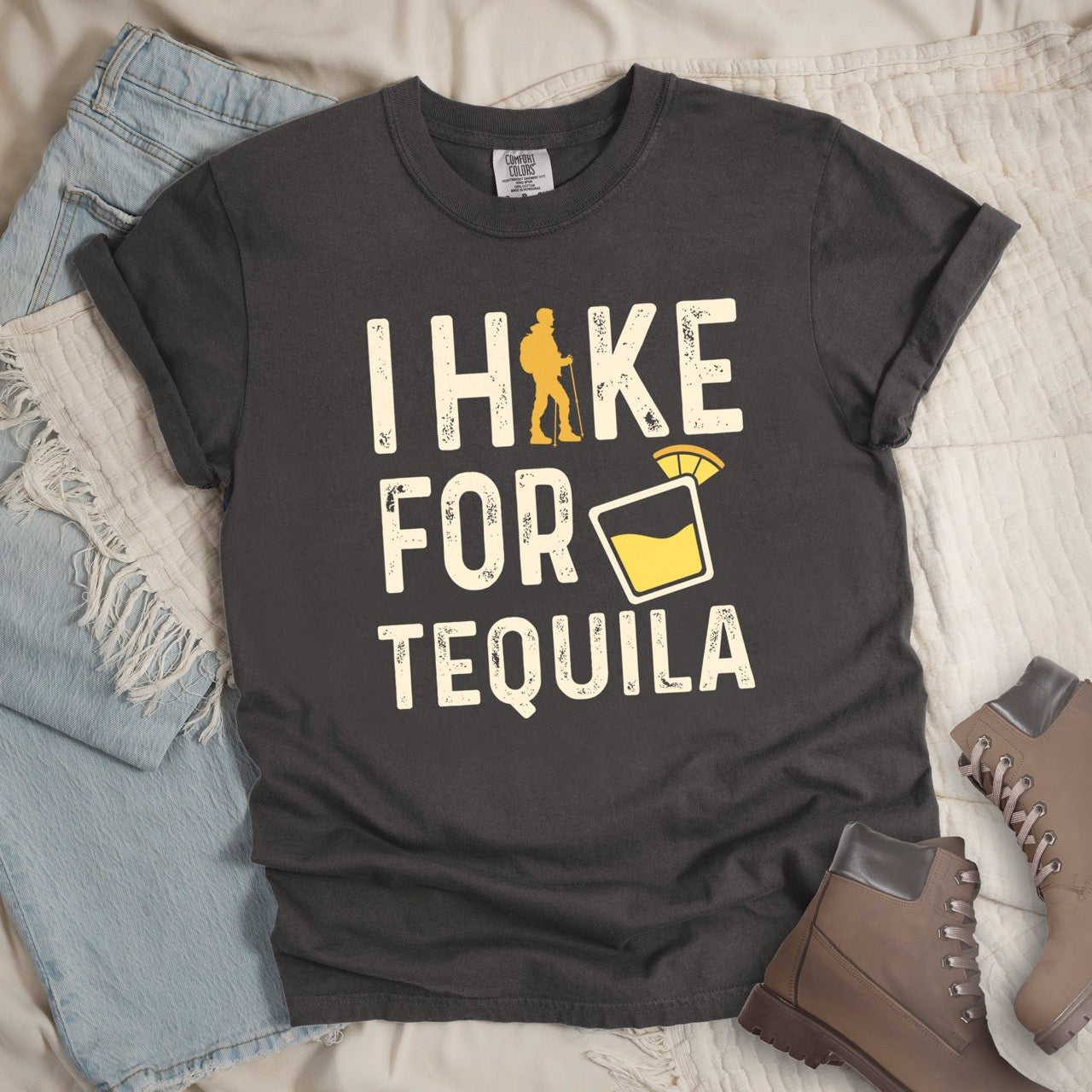 Pepper (Grey) Tequila margarita lover statement tee I Hike for Tequila with vintage distressed cream typography character silhouette with trekking pole and illustrated tilted margarita glass with lime garnish on Comfort Colors shirt for drink enthusiasts for hikers and foodies - laid flat with pants adn boots. 