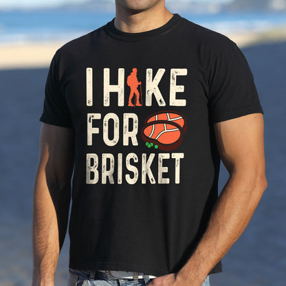 Black colored graphic t-shirt with 'I Hike for Brisket' text in white lettering featuring orange hiker silhouette and sliced brisket illustration with green garnish.