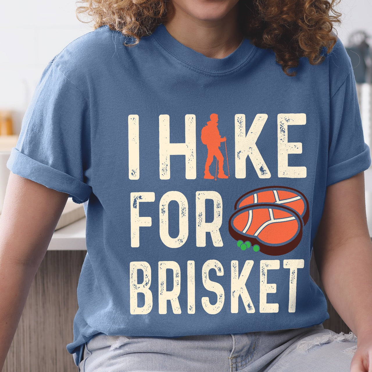 Woman wearing Blue Jean (blue) BBQ brisket lover statement tee I Hike for Brisket with distressed brown typography character silhouette with trekking pole and illustrated sliced smoked brisket with herb garnish on vintage Comfort Colors shirt for food motivated adventurers for hikers and foodies. 