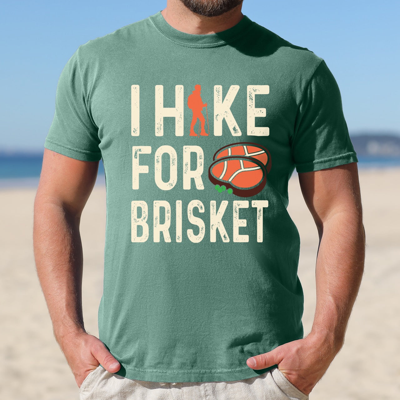 Man wearing Light Green BBQ brisket lover statement tee I Hike for Brisket with distressed brown typography character silhouette with trekking pole and illustrated sliced smoked brisket with herb garnish on vintage Comfort Colors shirt for food motivated adventurers for hikers and foodies. 