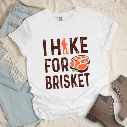 White colored graphic t-shirt with 'I Hike for Brisket' text in brown lettering featuring orange hiker silhouette and sliced brisket illustration with green garnish.