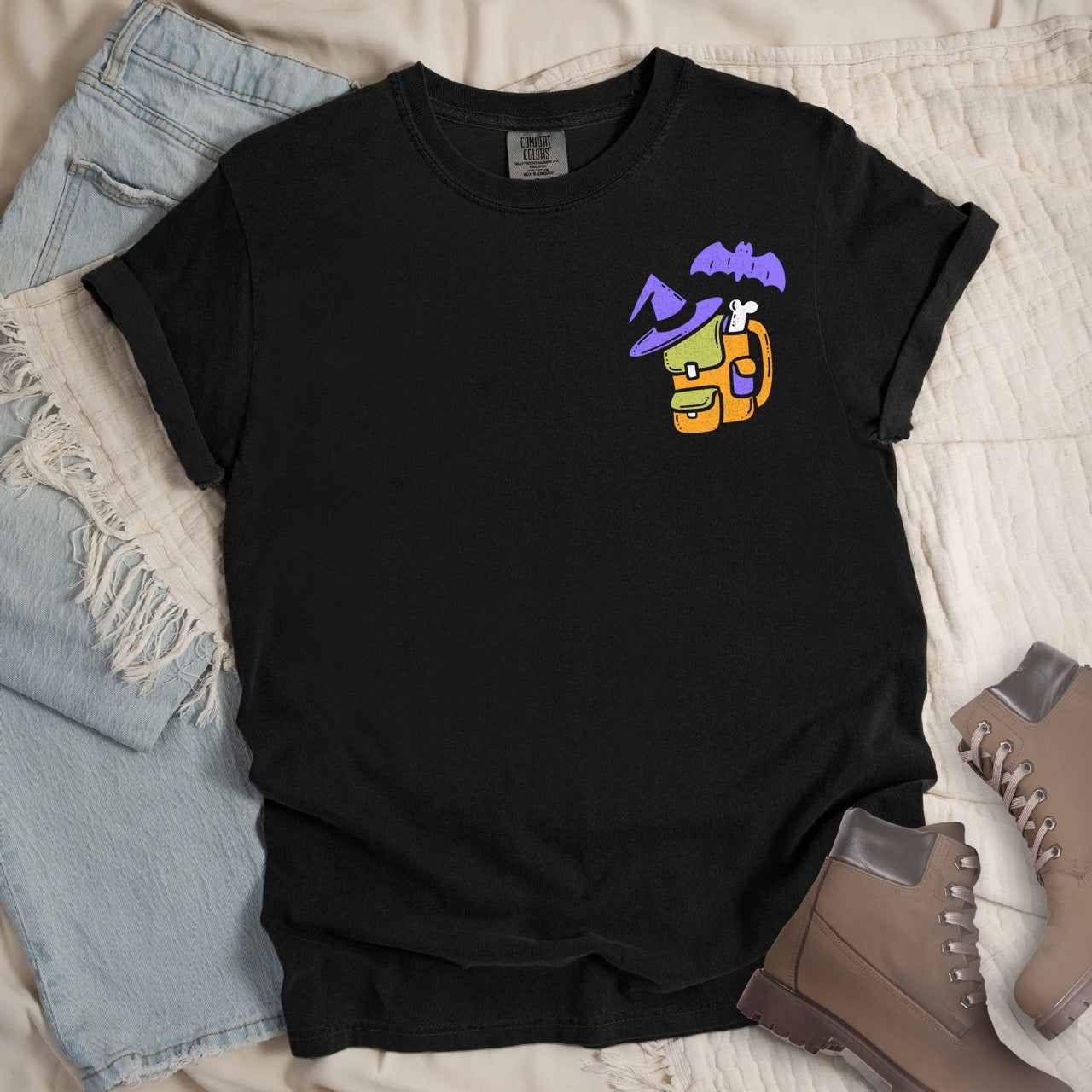 Black t-shirt with colorful graphic design  of a halloween themed back pack, witch hat and bat on a textured surface with jeans and boots.
