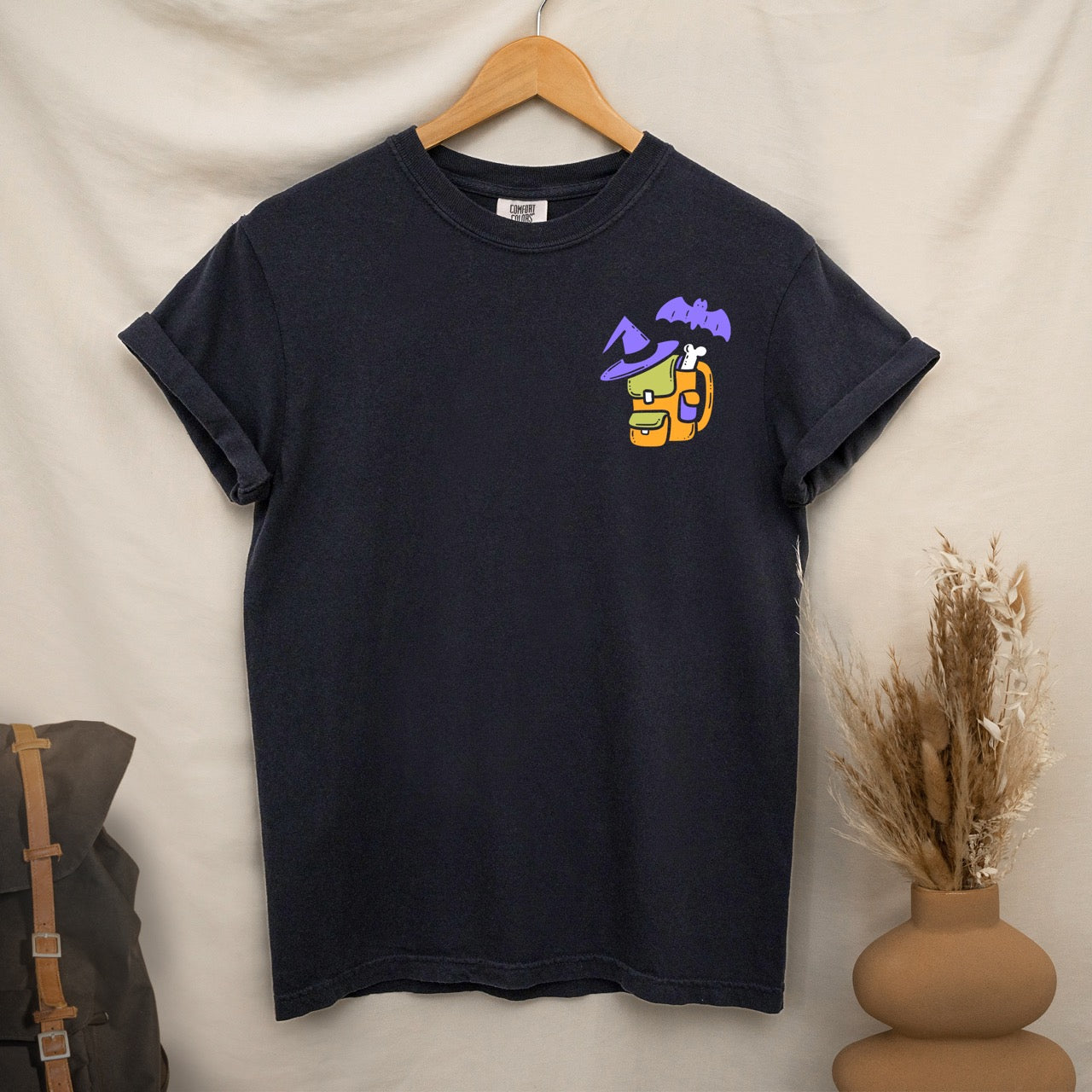 Black t-shirt with colorful graphic design  of a halloween themed back pack, witch hat and bat on a textured surface with jeans and boots hanging on a wooden hanger