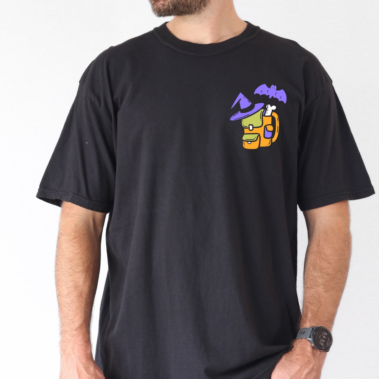 person wearing Black t-shirt with colorful graphic design  of a halloween themed back pack, witch hat and bat on a textured surface with jeans and boots.