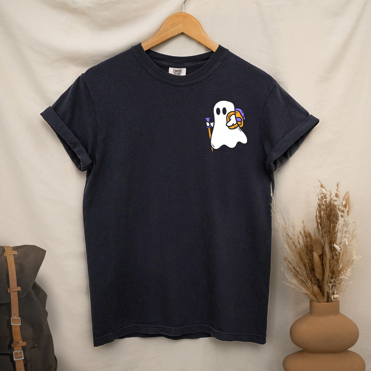 hanging  black Halloween ghost statement shirt featuring cute white Boo character with trekking pole and orange candy bucket filled with purple wrapped treats on chest placement Comfort Colors tee for spooky season celebrations for hikers and adventurers. 
