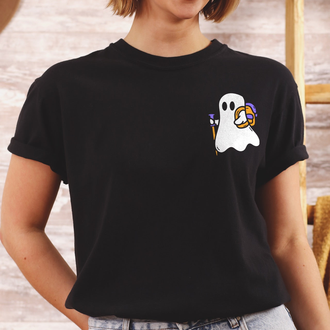 Woman wearing black Halloween ghost statement shirt featuring cute white Boo character with trekking pole and orange candy bucket filled with purple wrapped treats on chest placement Comfort Colors tee for spooky season celebrations for hikers and adventurers. 