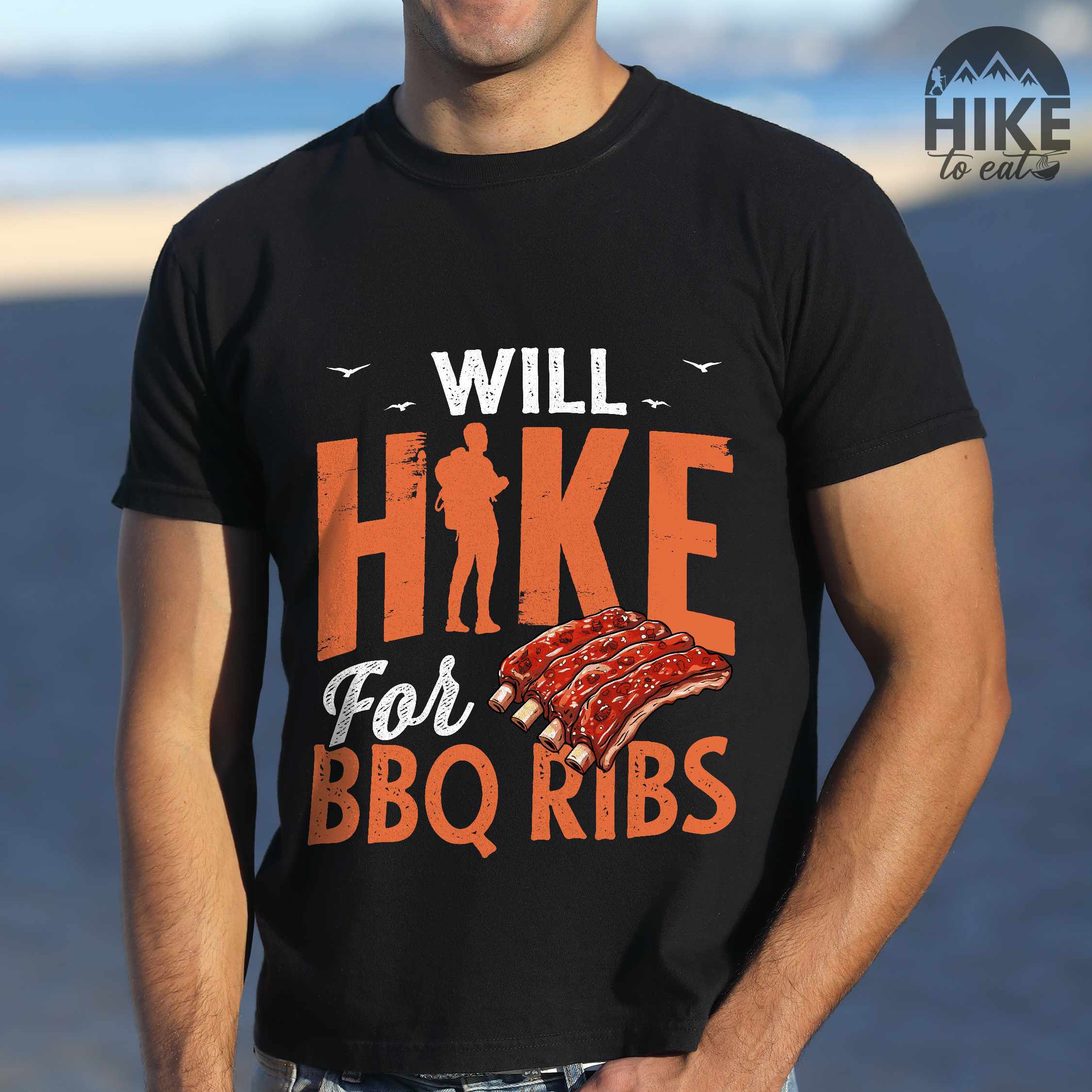 Man wearing black "Will Hike for BBQ Ribs" colored vintage tee with bold typography, birds, juicy grilled bbq ribs illustration, and backpacker for bbq enthusiasts