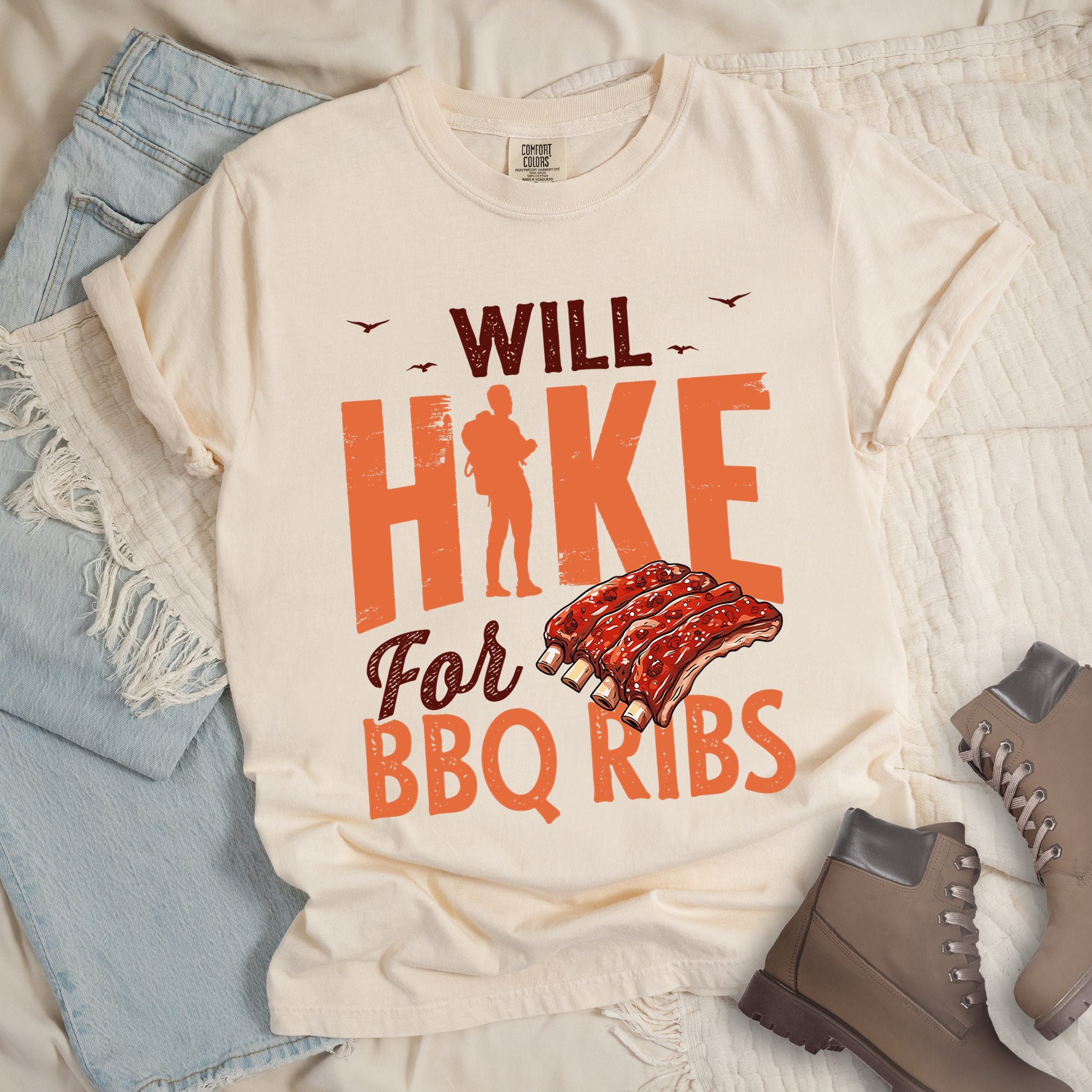 Ivory t-shirt with text "WILL HIKE For BBQ RIBS". "WILL" appears in deep brown-red distressed letters at the top with small bird silhouettes. "HIKE" is in large orange distressed text with a hiker silhouette replacing the letter "I". "For" appears in deep brown-red script, and "BBQ RIBS" in matching orange at the bottom. Between "For" and "BBQ RIBS" is an illustration of a rack of barbecue ribs with exposed bones.