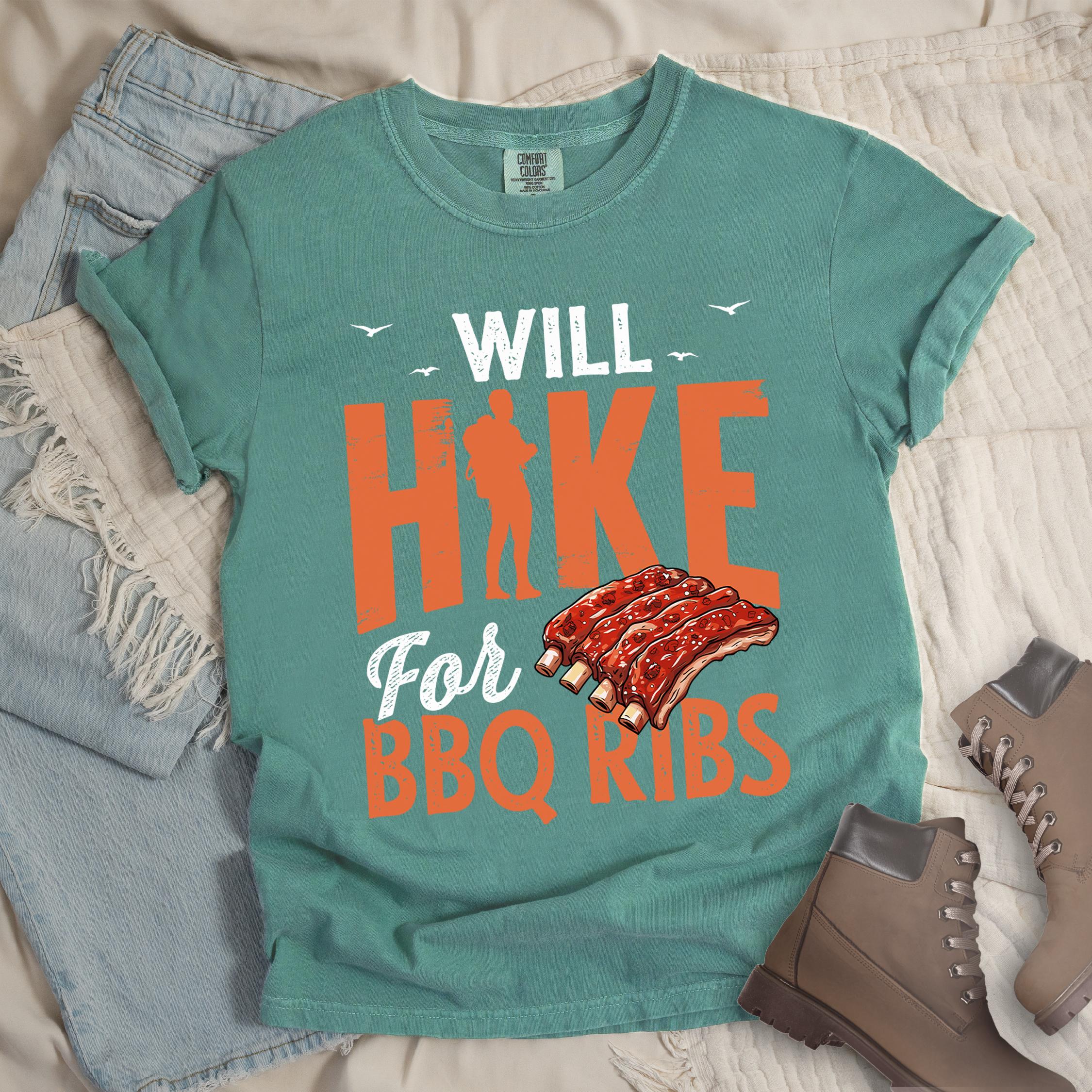 Green t-shirt with text "WILL HIKE For BBQ RIBS". "WILL" appears in deep brown-red distressed letters at the top with small bird silhouettes. "HIKE" is in large orange distressed text with a hiker silhouette replacing the letter "I". "For" appears in deep brown-red script, and "BBQ RIBS" in matching orange at the bottom. Between "For" and "BBQ RIBS" is an illustration of a rack of barbecue ribs with exposed bones.