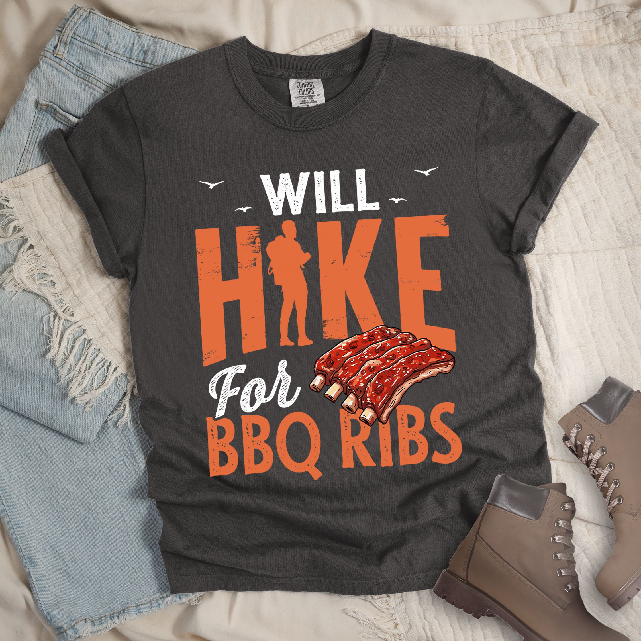 Dark grey t-shirt with text "WILL HIKE For BBQ RIBS". "WILL" appears in deep brown-red distressed letters at the top with small bird silhouettes. "HIKE" is in large orange distressed text with a hiker silhouette replacing the letter "I". "For" appears in deep brown-red script, and "BBQ RIBS" in matching orange at the bottom. Between "For" and "BBQ RIBS" is an illustration of a rack of barbecue ribs with exposed bones.