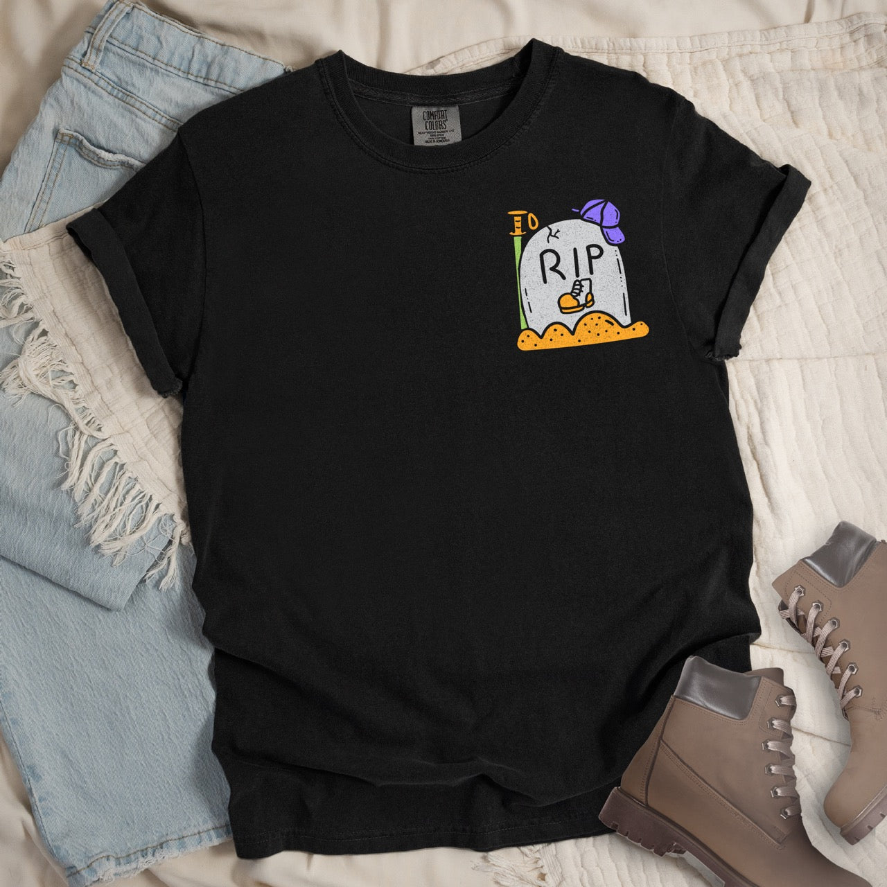 Black t-shirt with a colorful graphic design of a RIP boot on a textured surface with jeans and boots.