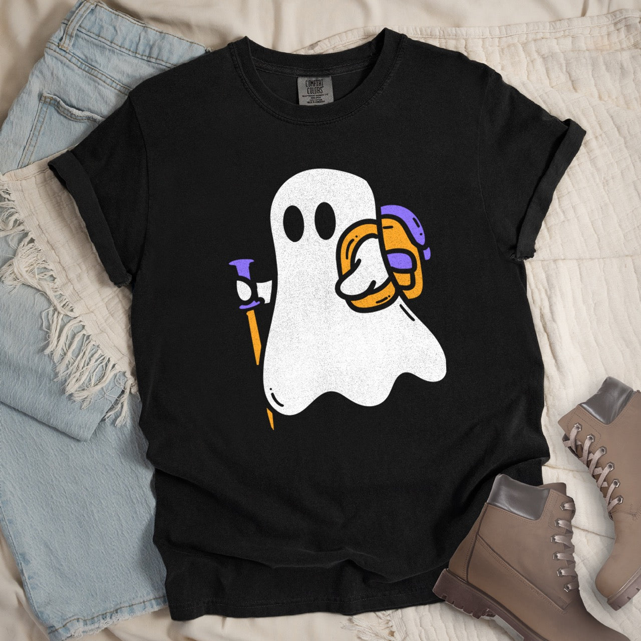 Black Halloween ghost statement tee with large full front design featuring cute white Boo character holding orange and purple trekking pole with colorful backpack perfect for spooky season celebrations on Comfort Colors shirt for hikers and adventurers - laid flt with boots and pants. 