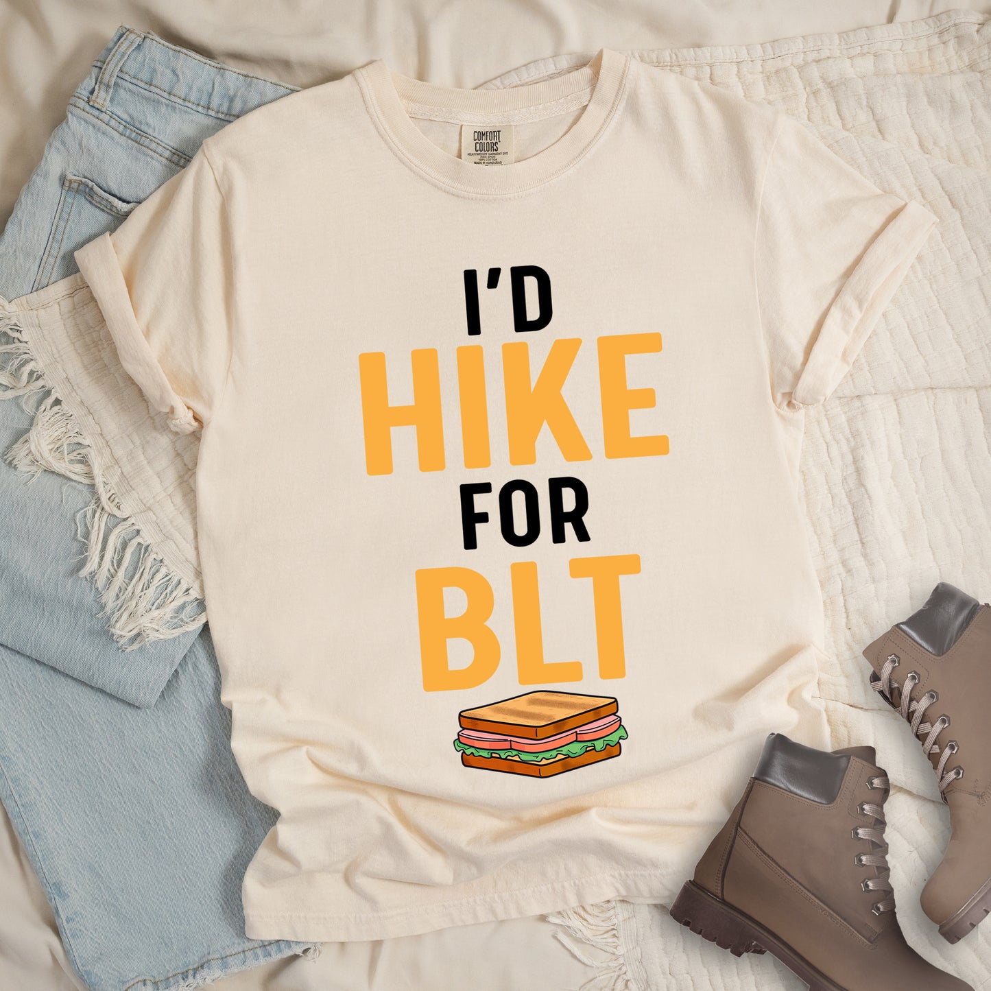 Ivory (Cream White) t-shirt with text "I'D HIKE FOR BLT". The words "I'D" appear in black at the top, "HIKE" in large gold/yellow letters in the middle, "FOR" in black, and "BLT" in matching gold/yellow at the bottom. Below the text is a colorful illustration of a BLT sandwich showing toast, maybe bacon, lettuce, and tomato layers. The simple, bold design emphasizes hiking motivation through food rewards.