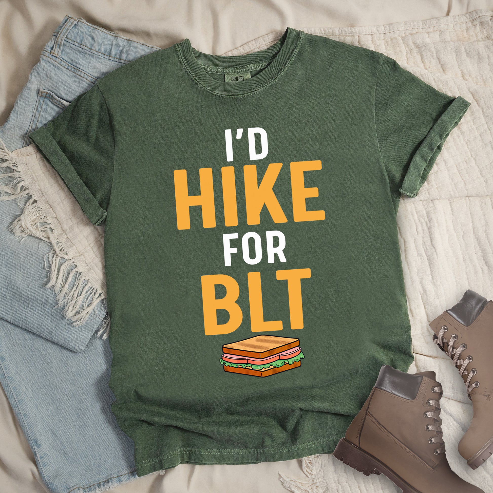 Moss (Dark Green) t-shirt with text "I'D HIKE FOR BLT". The words "I'D" appear in white at the top, "HIKE" in large gold/yellow letters in the middle, "FOR" in white, and "BLT" in matching gold/yellow at the bottom. Below the text is a colorful illustration of a BLT sandwich showing toast, maybe bacon, lettuce, and tomato layers. The simple, bold design emphasizes hiking motivation through food rewards.