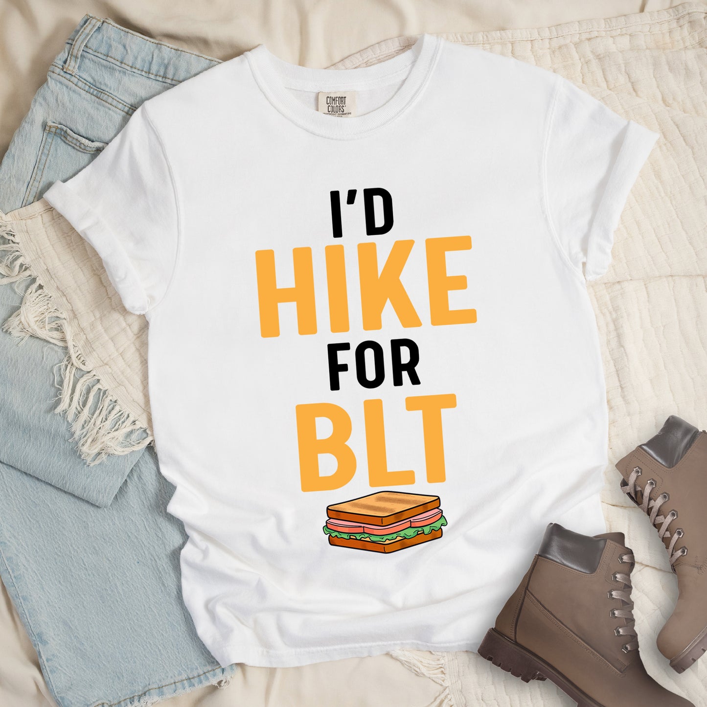 White t-shirt with text "I'D HIKE FOR BLT". The words "I'D" appear in black at the top, "HIKE" in large gold/yellow letters in the middle, "FOR" in black, and "BLT" in matching gold/yellow at the bottom. Below the text is a colorful illustration of a BLT sandwich showing toast, maybe bacon, lettuce, and tomato layers. The simple, bold design emphasizes hiking motivation through food rewards.