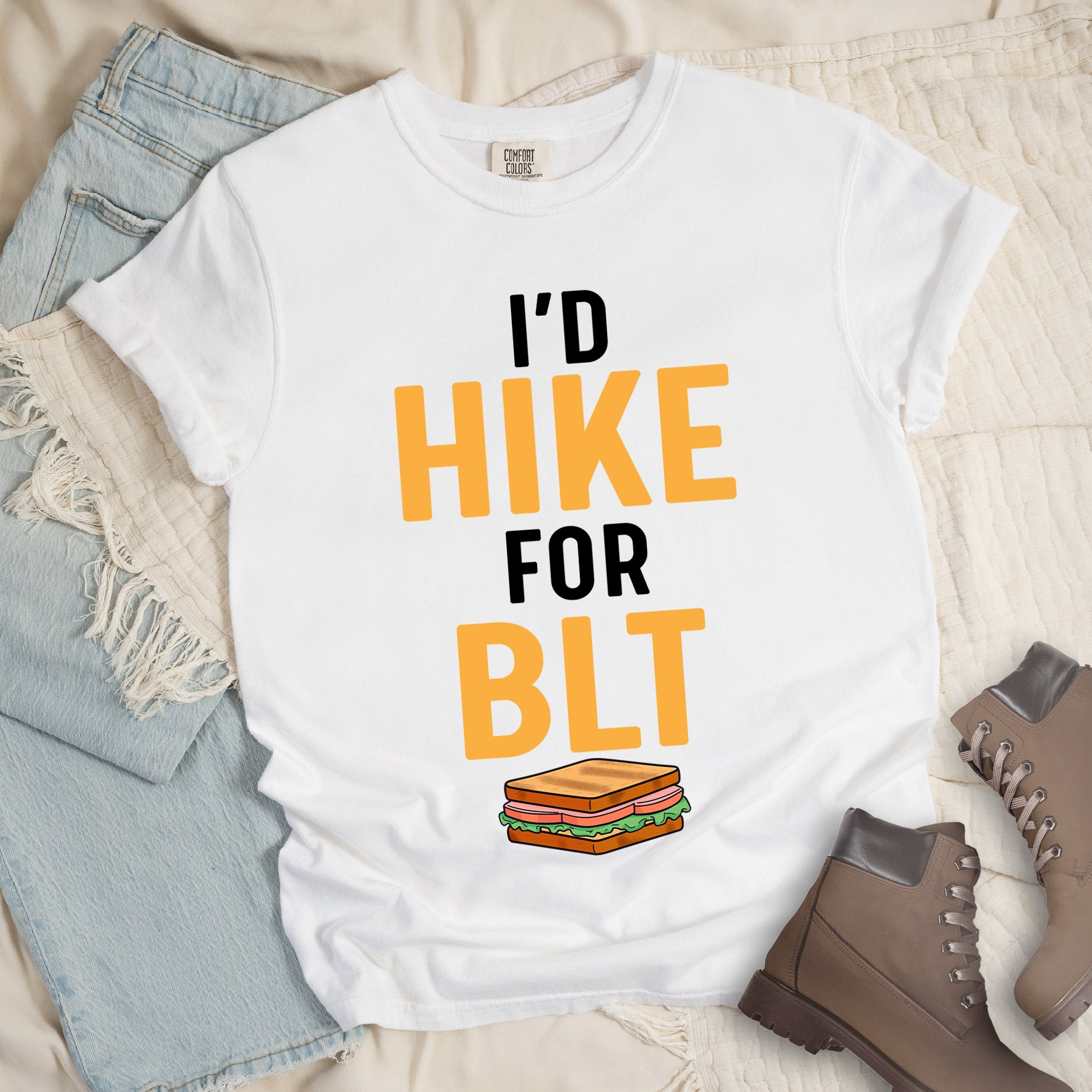 White t-shirt with text "I'D HIKE FOR BLT". The words "I'D" appear in white at the top, "HIKE" in large gold/yellow letters in the middle, "FOR" in white, and "BLT" in matching gold/yellow at the bottom. Below the text is a colorful illustration of a BLT sandwich showing toast, maybe bacon, lettuce, and tomato layers. The simple, bold design emphasizes hiking motivation through food rewards.