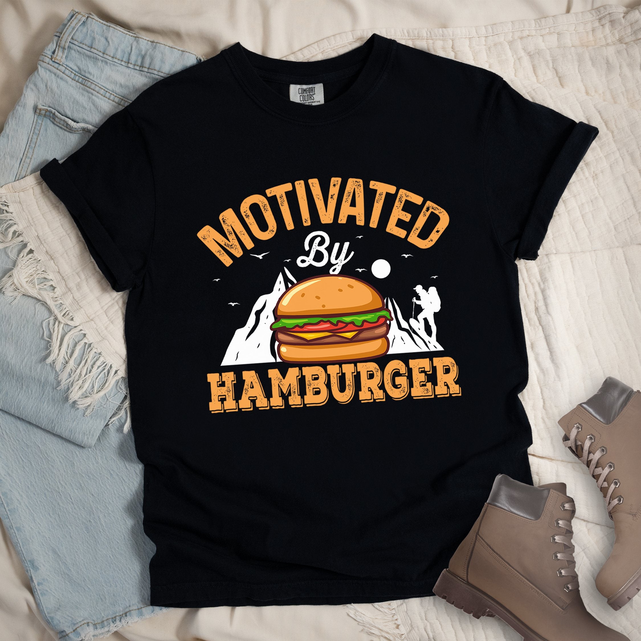 Black t-shirt with text "MOTIVATED By HAMBURGER". The word "MOTIVATED" appears in orange distressed letters at the top in an arc, "By" in white script in the middle, and "HAMBURGER" in matching orange at the bottom. The design features white mountain silhouettes with a small hiker figure, flying birds, and a sun or moon. A colorful hamburger illustration with bun, patty, cheese, lettuce and tomato sits in the center of the design.