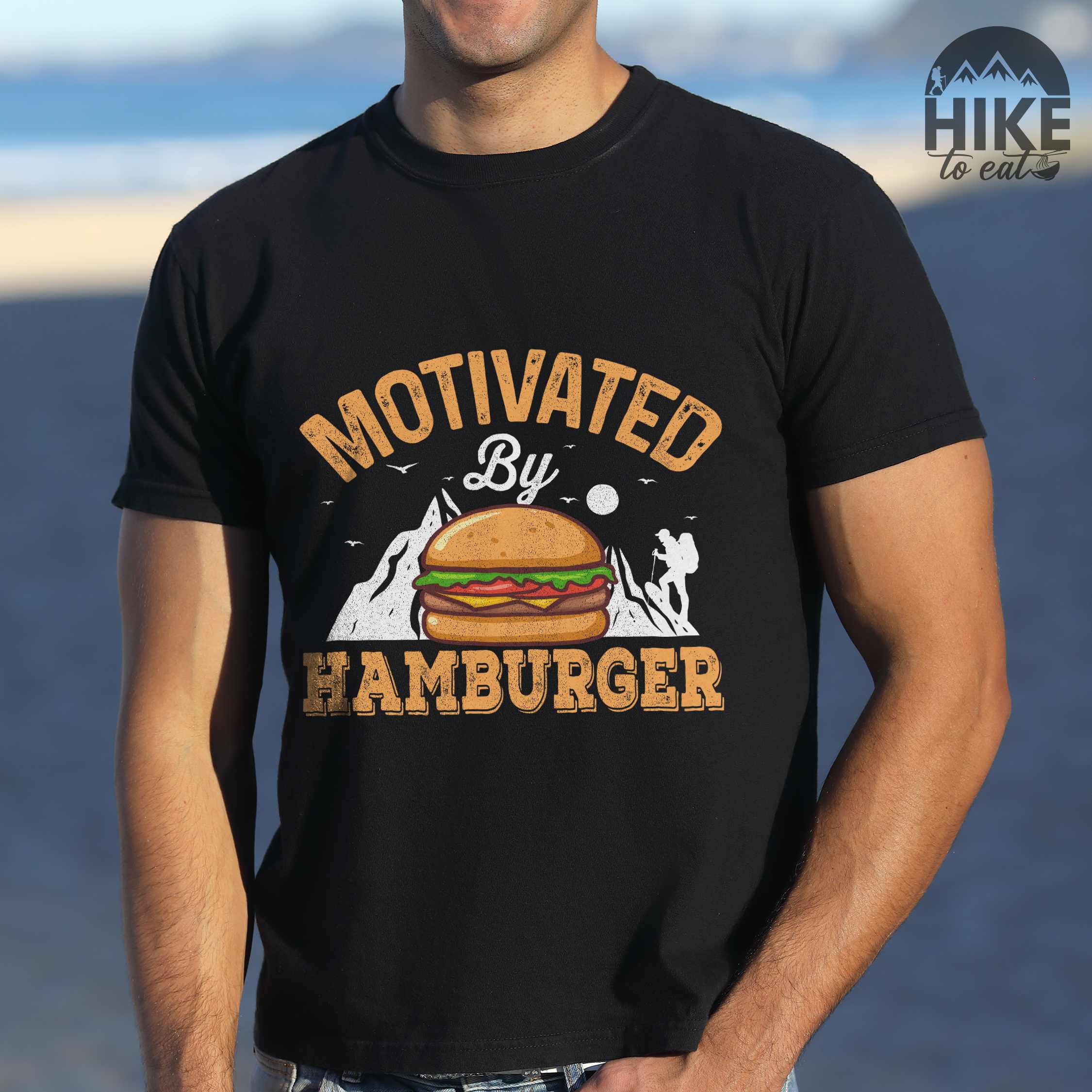Man wearing black "Motivated by Hamburger" colored vintage tee with bold typography, birds, mountains, illustrated juicy hamburger, and backpacker for burger enthusiasts.