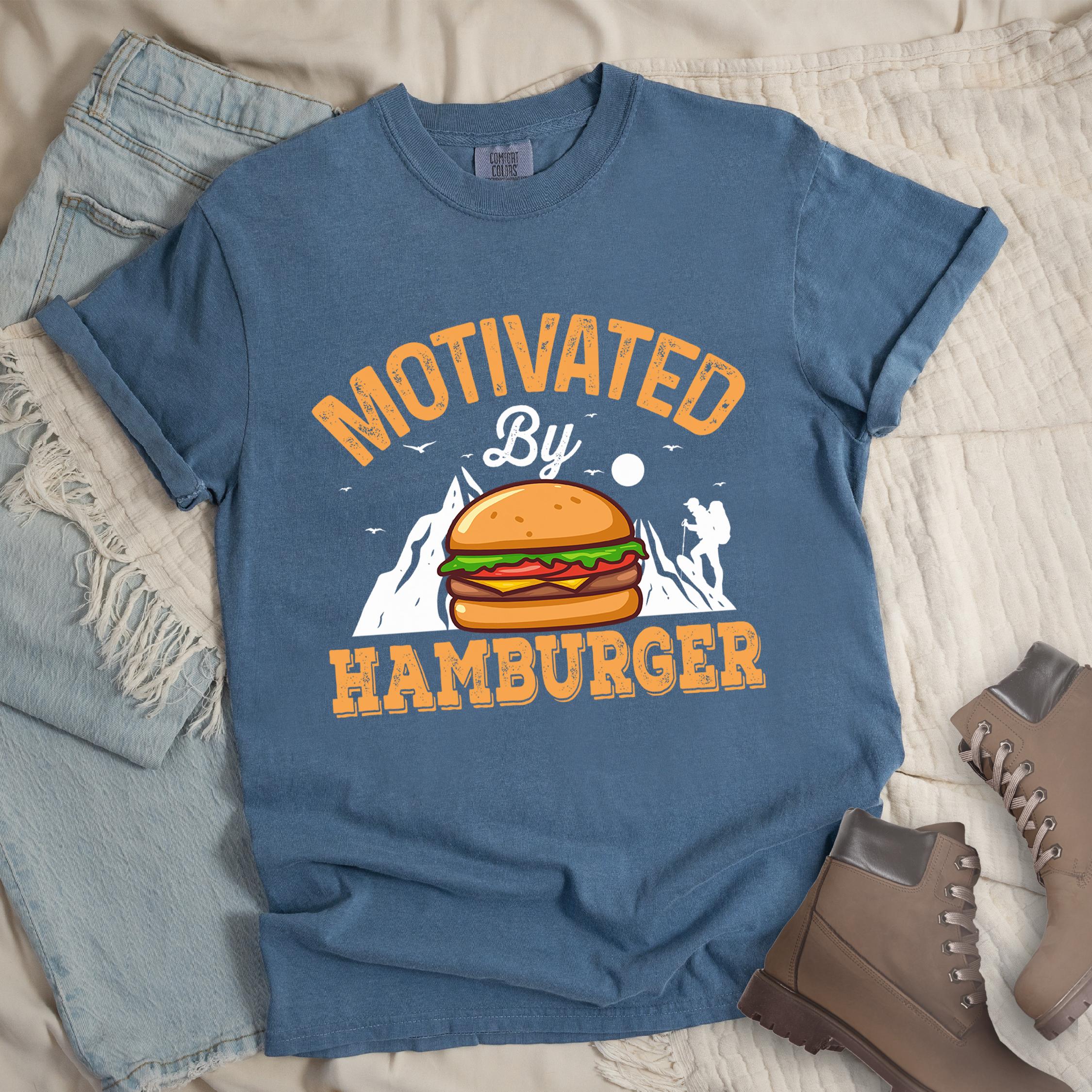 Dark blue t-shirt with text "MOTIVATED By HAMBURGER". The word "MOTIVATED" appears in distressed letters at the top in an arc, "By" in script in the middle, and "HAMBURGER" in matching orange at the bottom. The design features white mountain silhouettes with a small hiker figure, flying birds, and a sun or moon. A colorful hamburger illustration with bun, patty, cheese, lettuce and tomato sits in the center of the design.