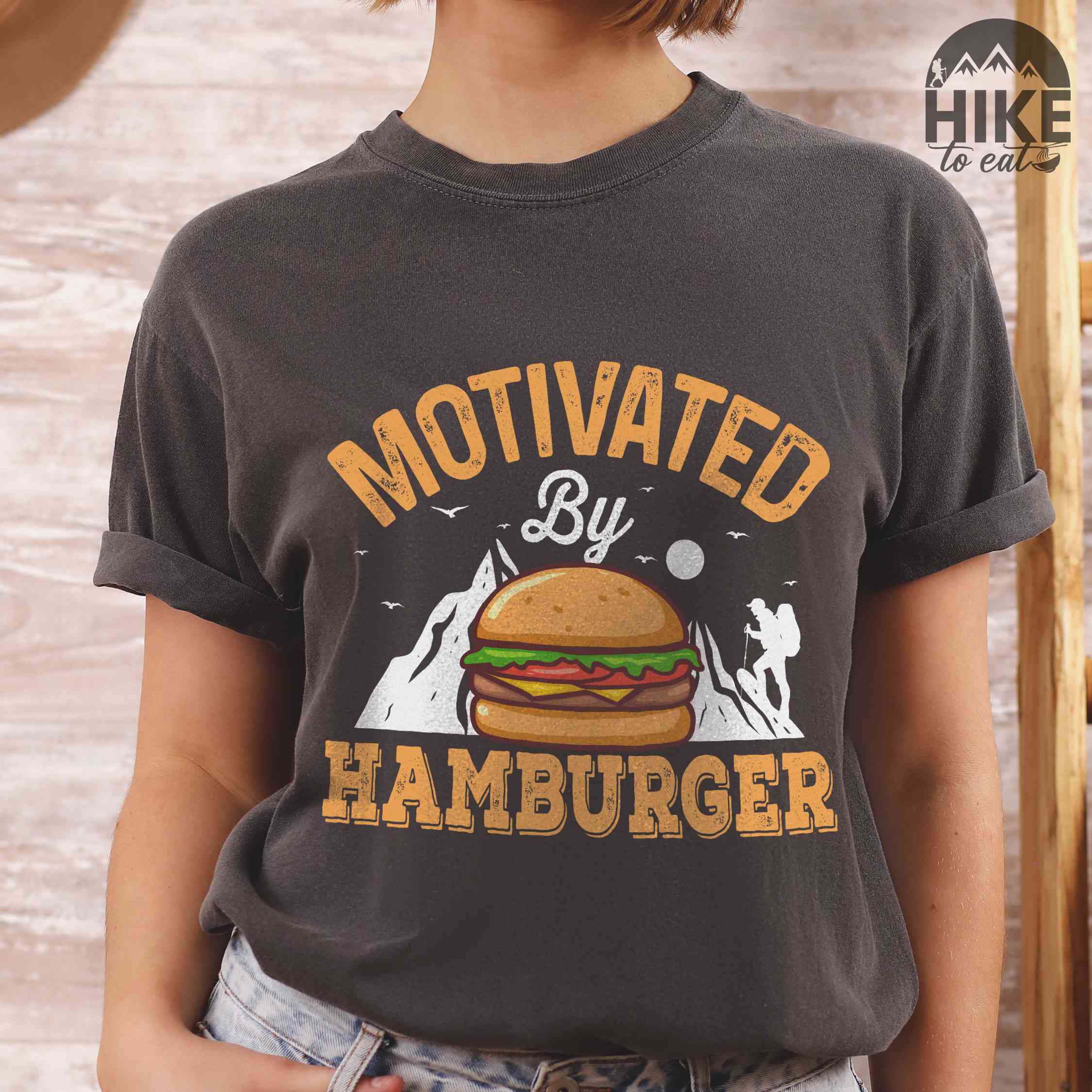Woman wearing pepper "Motivated by Hamburger" colored vintage tee with bold typography, birds, mountains, illustrated juicy hamburger, and backpacker for burger enthusiasts.