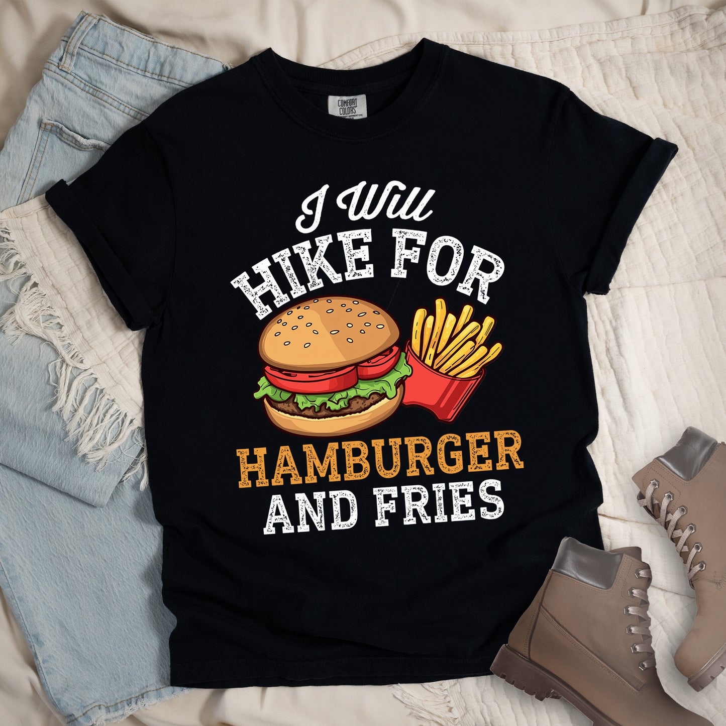Black t-shirt with white distressed text reading "I Will HIKE FOR HAMBURGER AND FRIES". The words "I Will" appear in white script at the top, "HIKE FOR" in white block letters, "HAMBURGER" in gold/orange, and "AND FRIES" in white at the bottom. Center illustration shows a detailed hamburger with bun, patty, lettuce, and tomato next to a red container of yellow french fries.