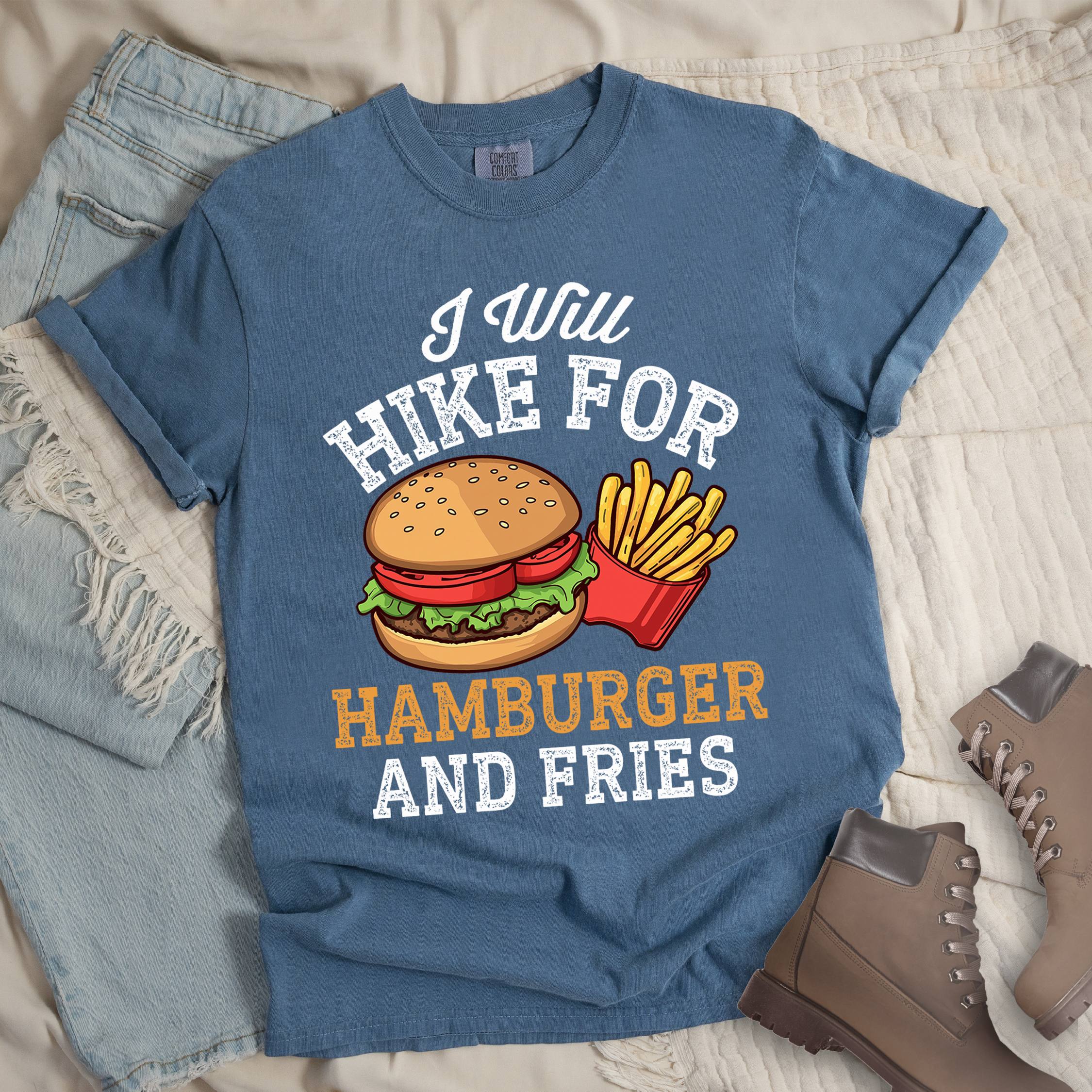 Dark blue t-shirt with distressed text reading "I Will HIKE FOR HAMBURGER AND FRIES". The words "I Will" appear in script at the top, "HIKE FOR" in white block letters, "HAMBURGER" in gold/orange, and "AND FRIES" in white at the bottom. Center illustration shows a detailed hamburger with bun, patty, lettuce, and tomato next to a red container of yellow french fries.