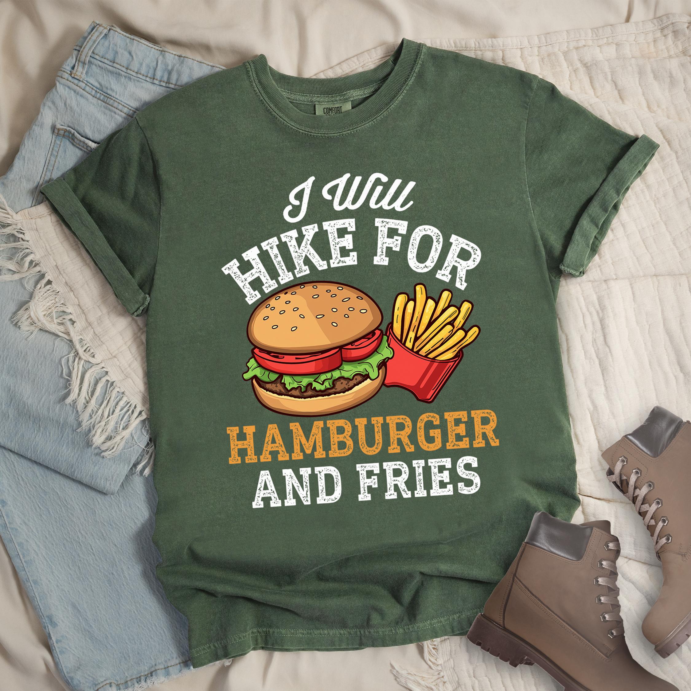 Dark green t-shirt with distressed text reading "I Will HIKE FOR HAMBURGER AND FRIES". The words "I Will" appear in script at the top, "HIKE FOR" in white block letters, "HAMBURGER" in gold/orange, and "AND FRIES" in white at the bottom. Center illustration shows a detailed hamburger with bun, patty, lettuce, and tomato next to a red container of yellow french fries.