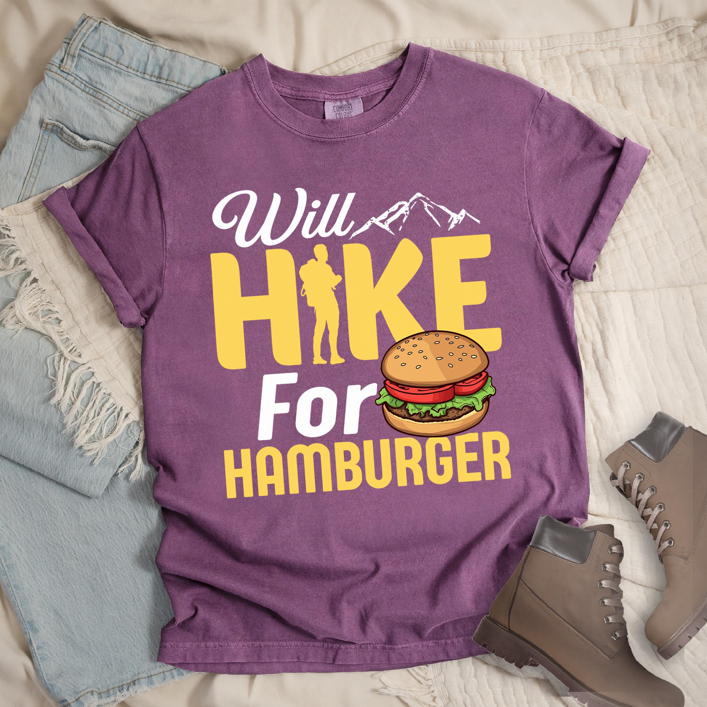 Dark pink t-shirt with text "Will HIKE For HAMBURGER". "Will" appears in white script with small mountain silhouettes. "HIKE" is in large yellow letters with a hiker silhouette replacing the letter "I". "For" text, and "HAMBURGER" at the bottom. Next to "For" and above "HAMBURGER" is a colorful illustration of a hamburger with sesame seed bun, lettuce, tomato, and patty.Features a classic crew neck and casual rolled sleeves, styled alongside light wash denim and brown hiking boots