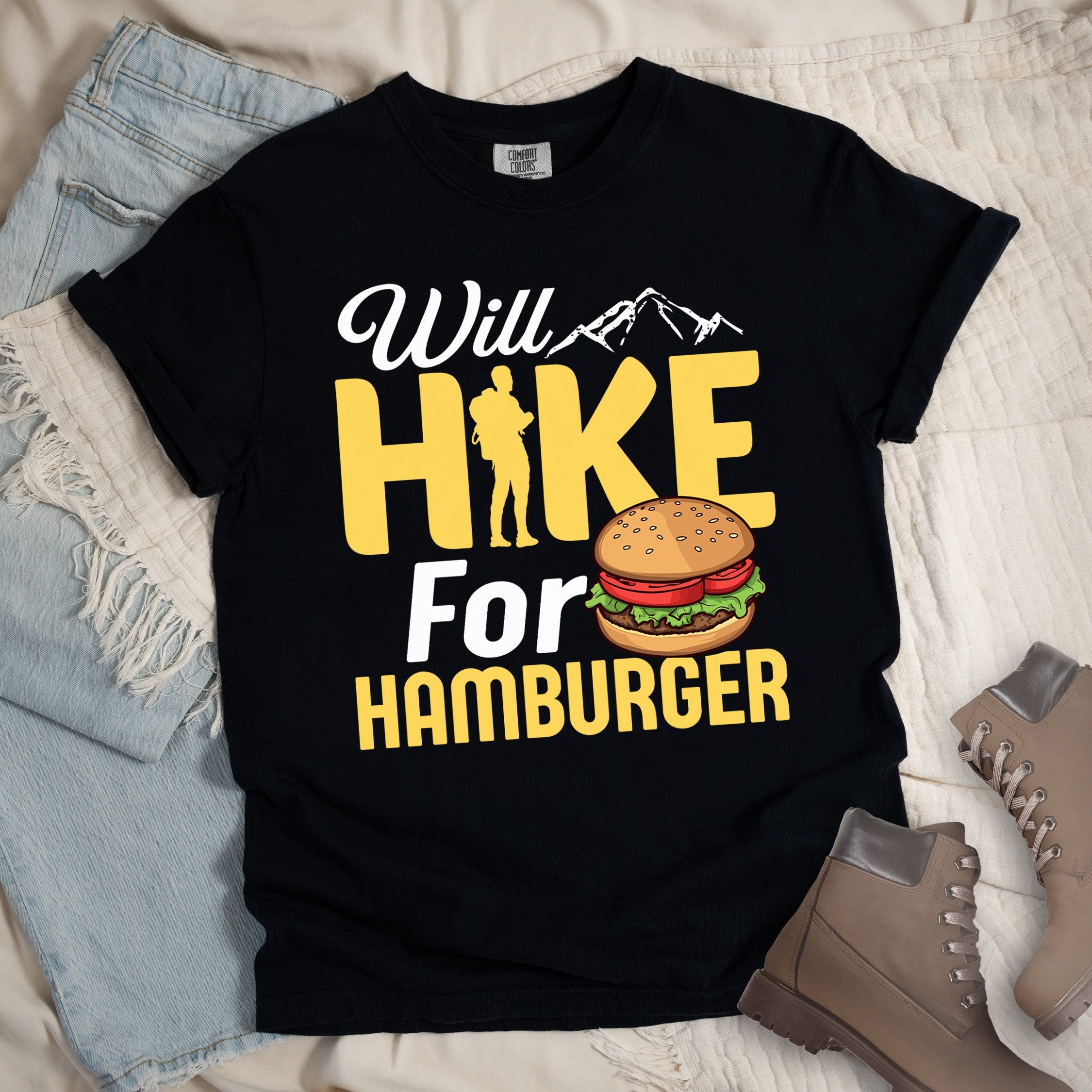 Black t-shirt with text "Will HIKE For HAMBURGER". "Will" appears in white script with small mountain silhouettes. "HIKE" is in large yellow letters with a hiker silhouette replacing the letter "I". "For" text, and "HAMBURGER" at the bottom. Next to "For" and above "HAMBURGER" is a colorful illustration of a hamburger with sesame seed bun, lettuce, tomato, and patty.Features a classic crew neck and casual rolled sleeves, styled alongside light wash denim and brown hiking boots