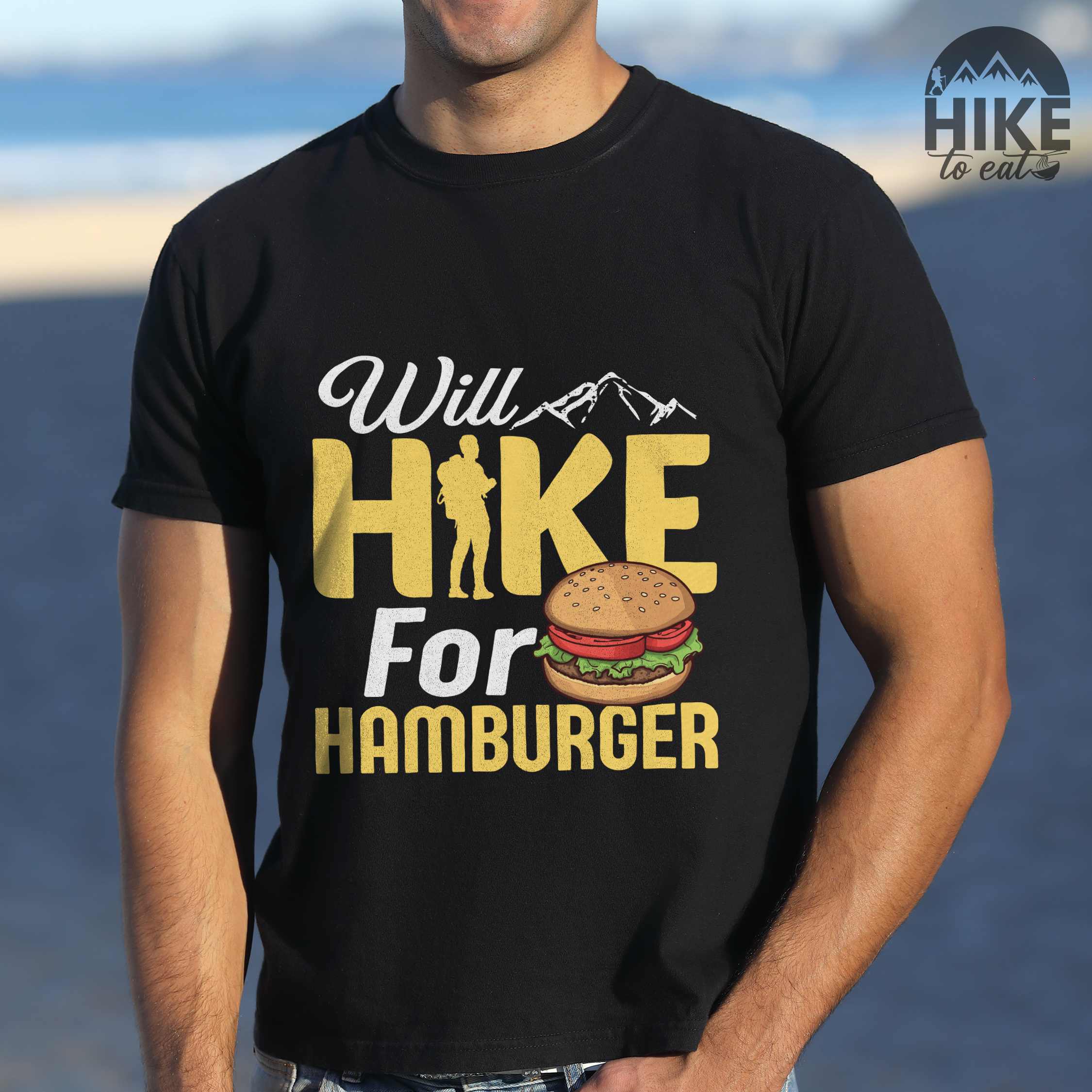 Man wearing black "Will Hike for Hamburger" colored vintage tee with bold typography, birds, mountains, illustrated juicy hamburger, and backpacker for burger enthusiasts.